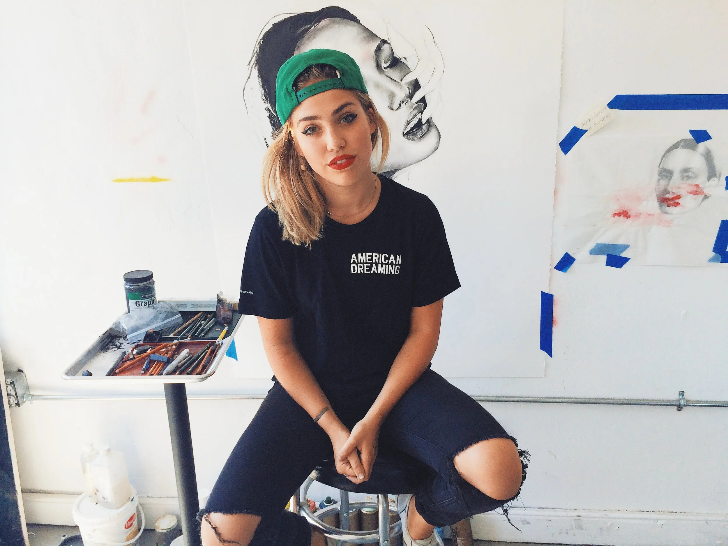 A young woman with blonde hair, wearing a green cap and a black t-shirt that says 'American Dreaming', sitting on a stool in front of a wall with art. There are art supplies on a small table next to her.
