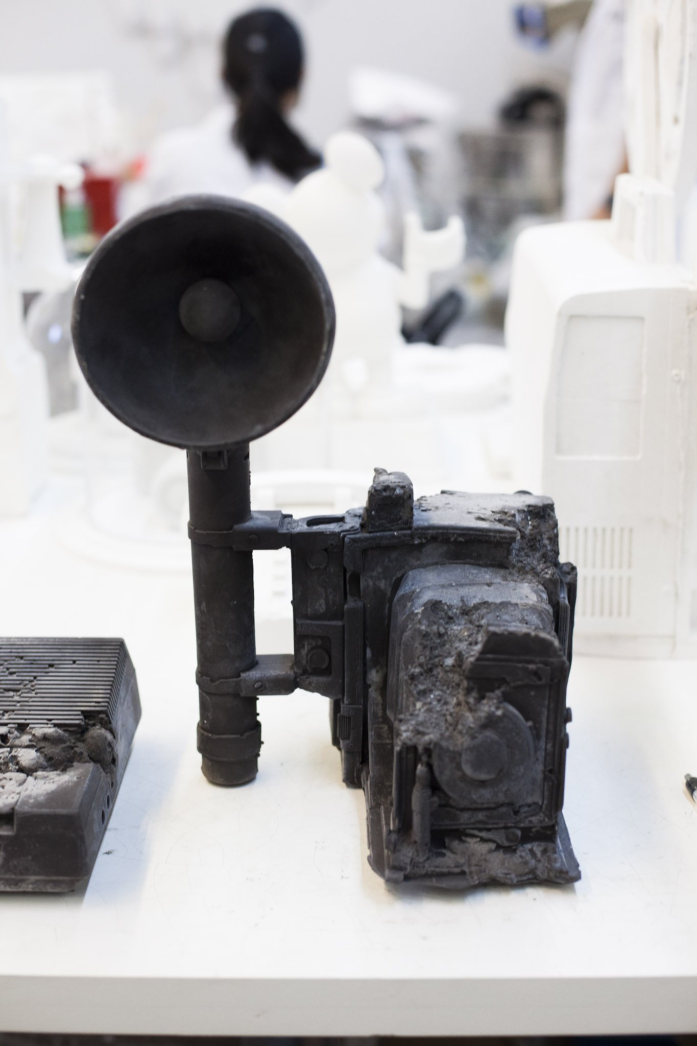 A burnt-out, charred camera on a white table with a blurred person working in the background.