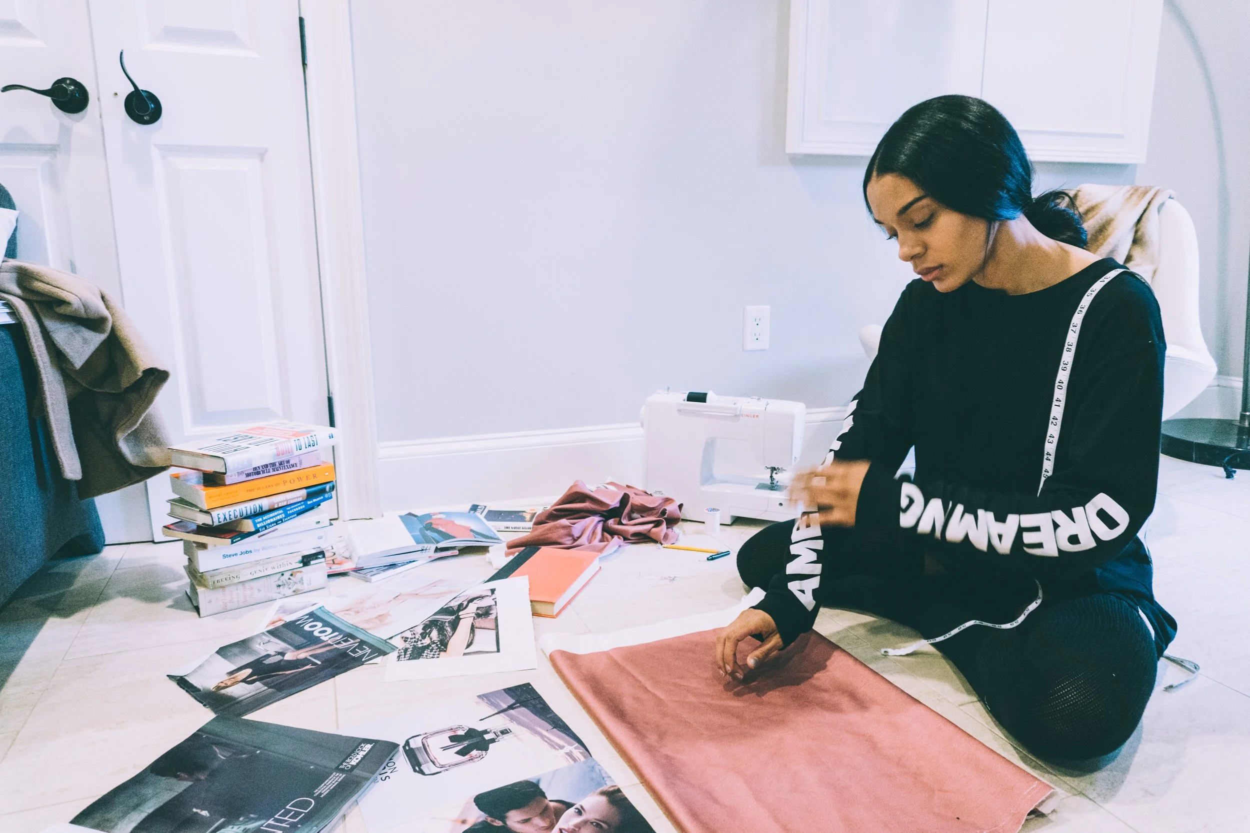A woman sitting on the floor surrounded by books, magazines, fabric, and sewing supplies, working on a sewing project.