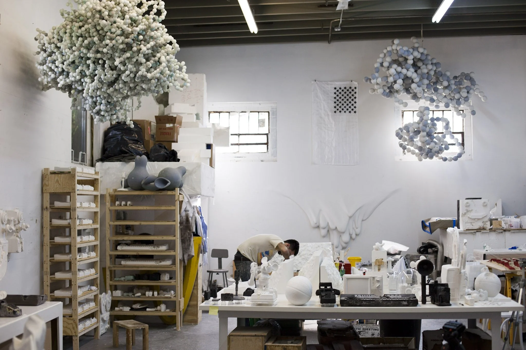 Artist's studio with various sculptures, art supplies, and artworks including cloud-like installations hanging from the ceiling and a wall sculpture of wings.