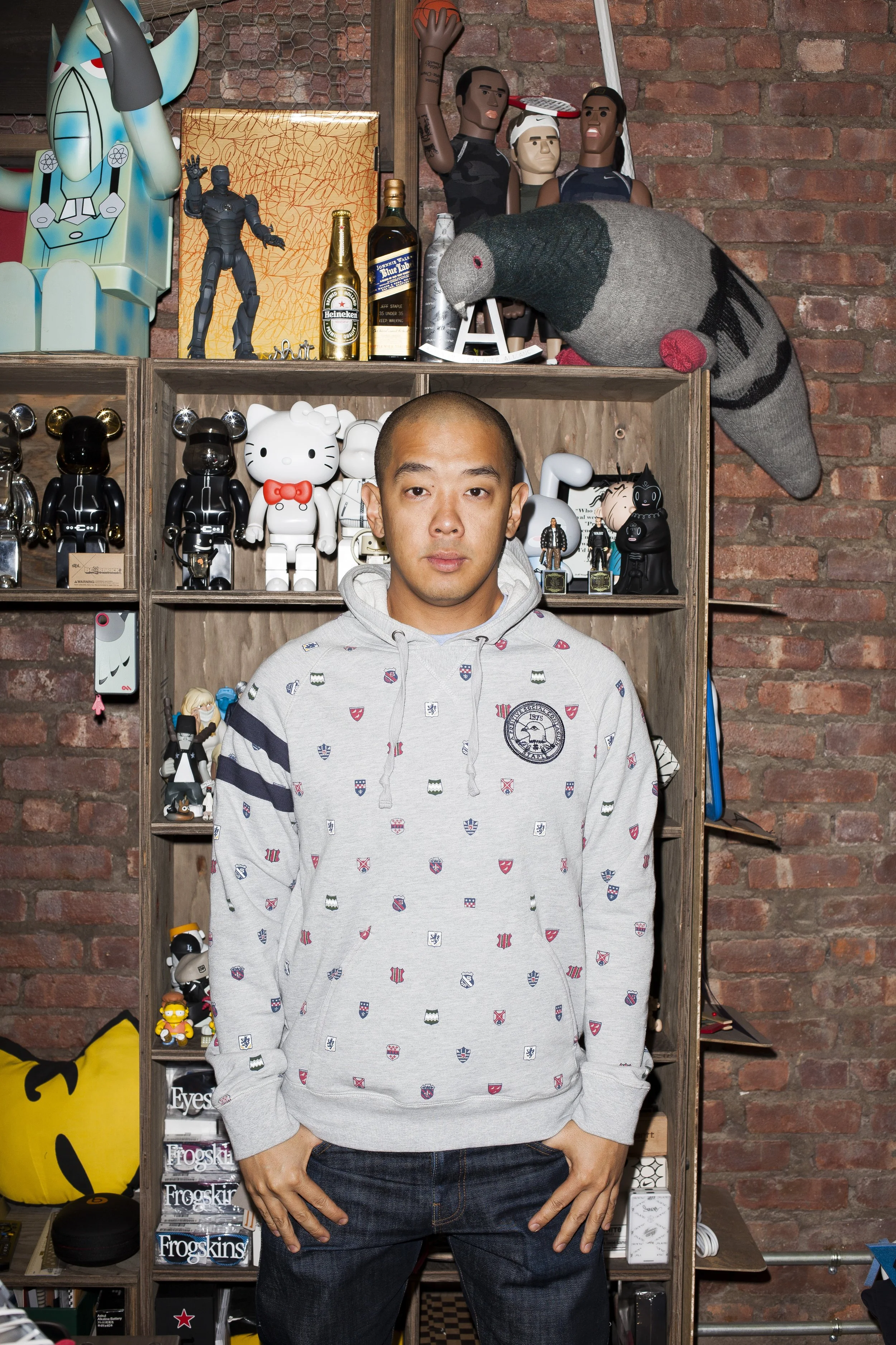 A man standing in front of a wooden shelf filled with collectible toys and art figures, wearing a gray hoodie with colorful patches, against a brick wall background.