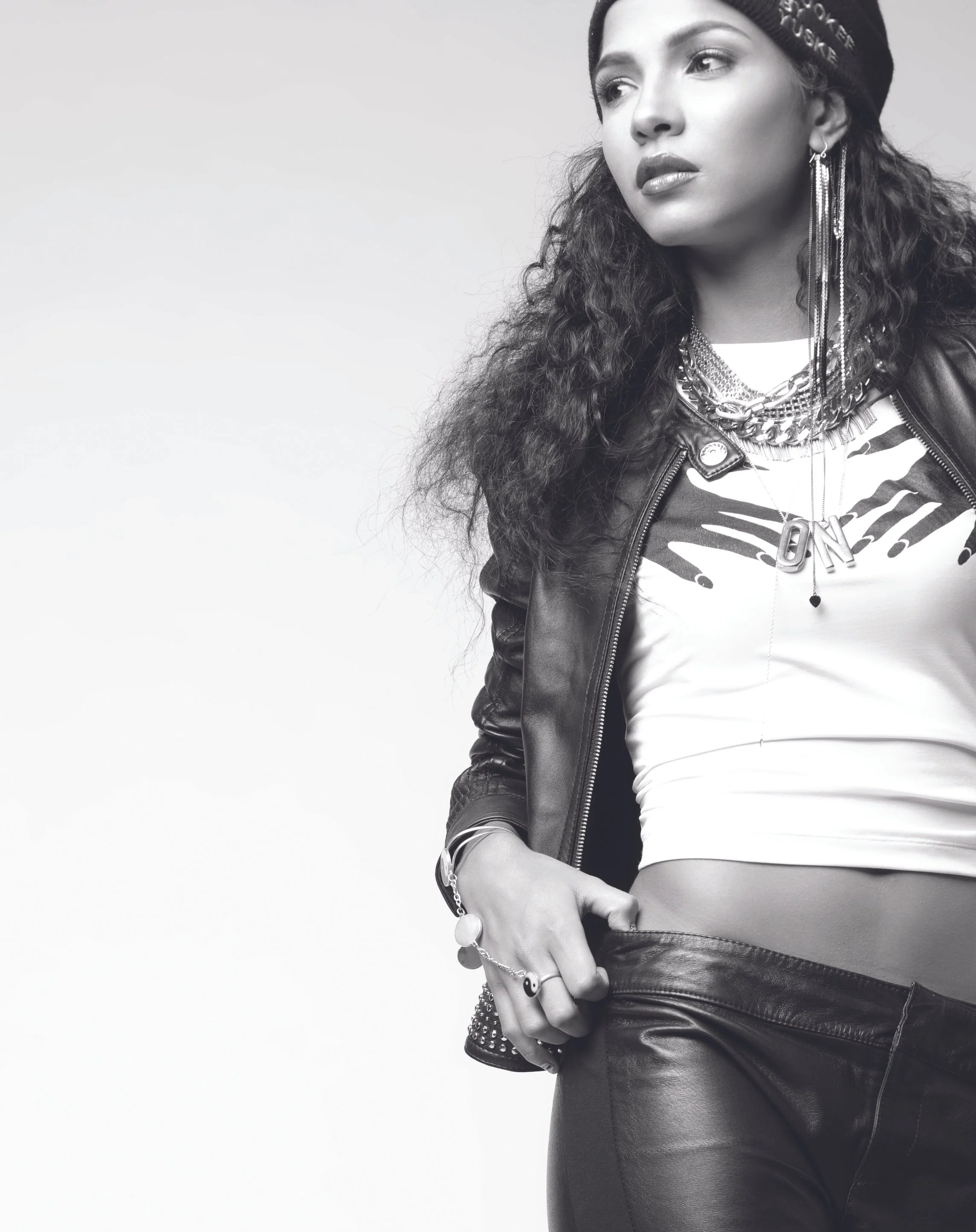 Black and white photo of a woman with curly hair wearing a black beanie, layered necklaces, earrings, a leather jacket, a graphic t-shirt, and leather pants.