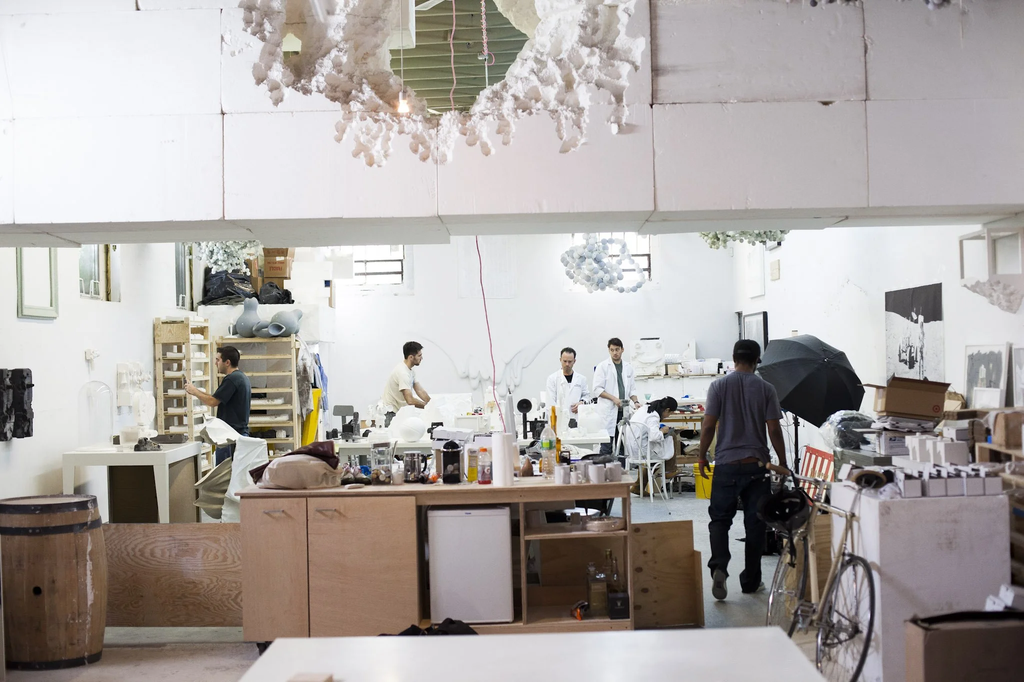 A spacious art studio with people working at tables surrounded by art supplies and materials. The studio has white walls, high ceilings, and large windows letting in natural light. There is a large decorative white paper or fabric piece hanging from the ceiling.