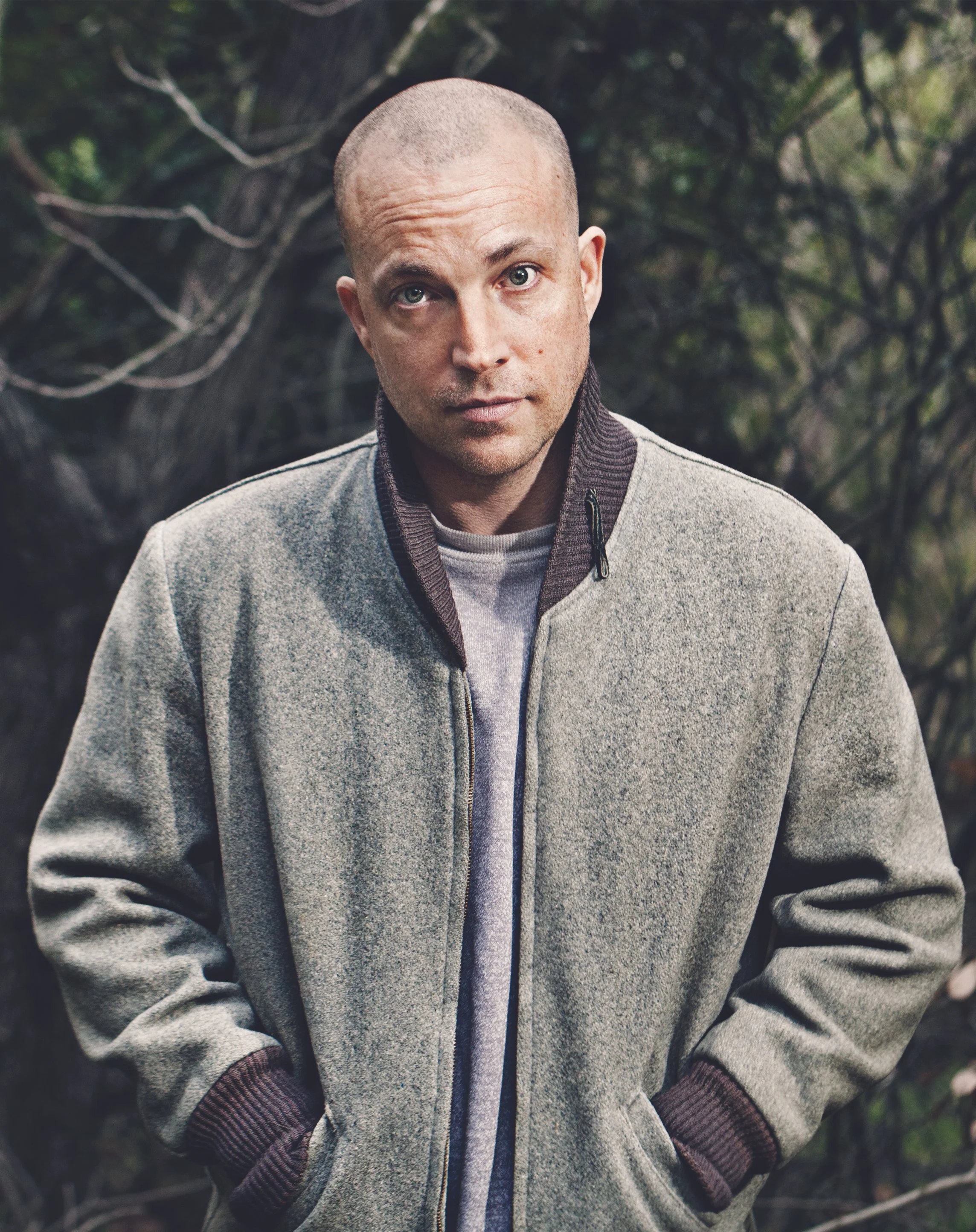 Man with a shaved head and casual gray jacket standing outdoors with trees in the background.