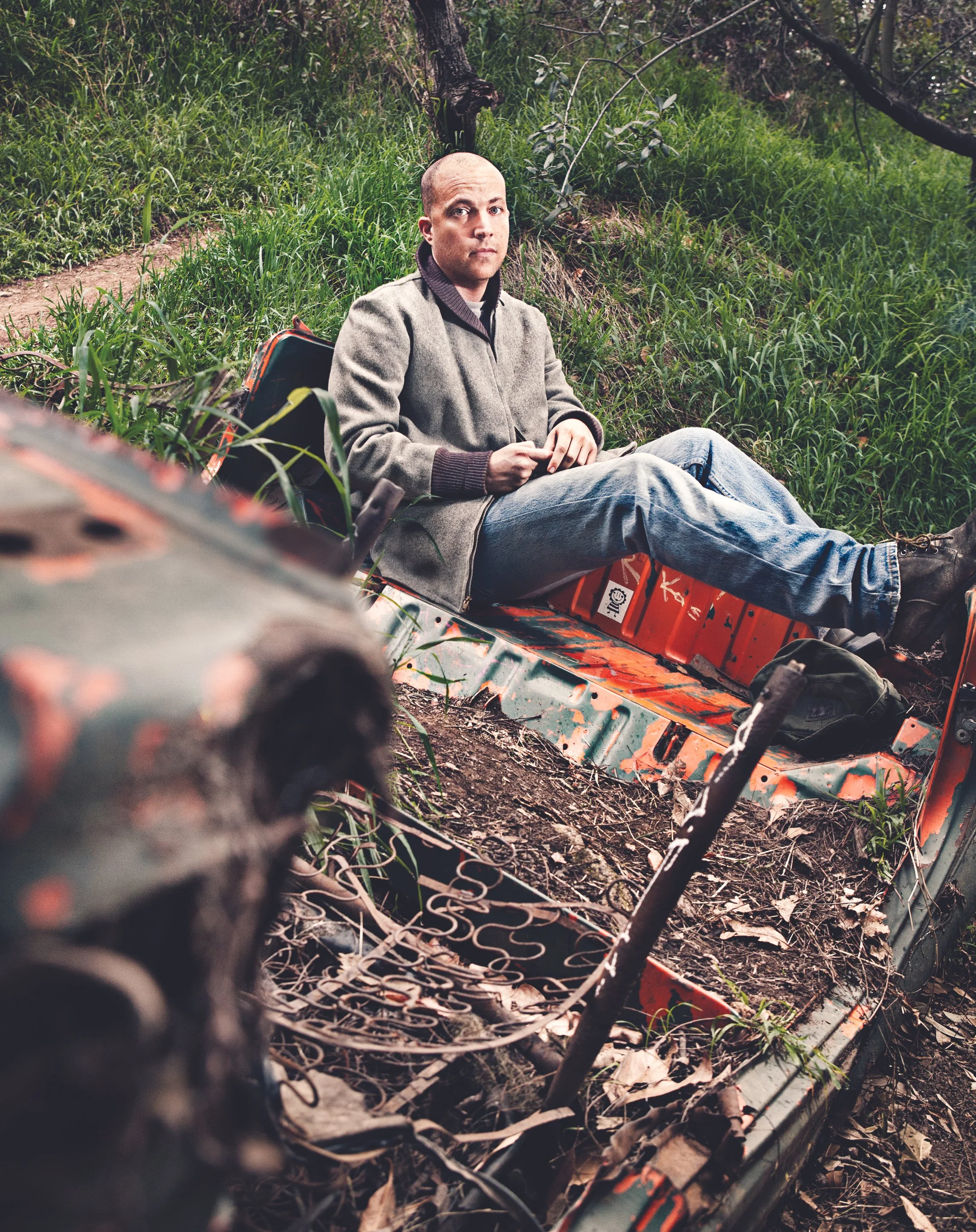 A man sitting in an overturned orange boat in a grassy, wooded area.