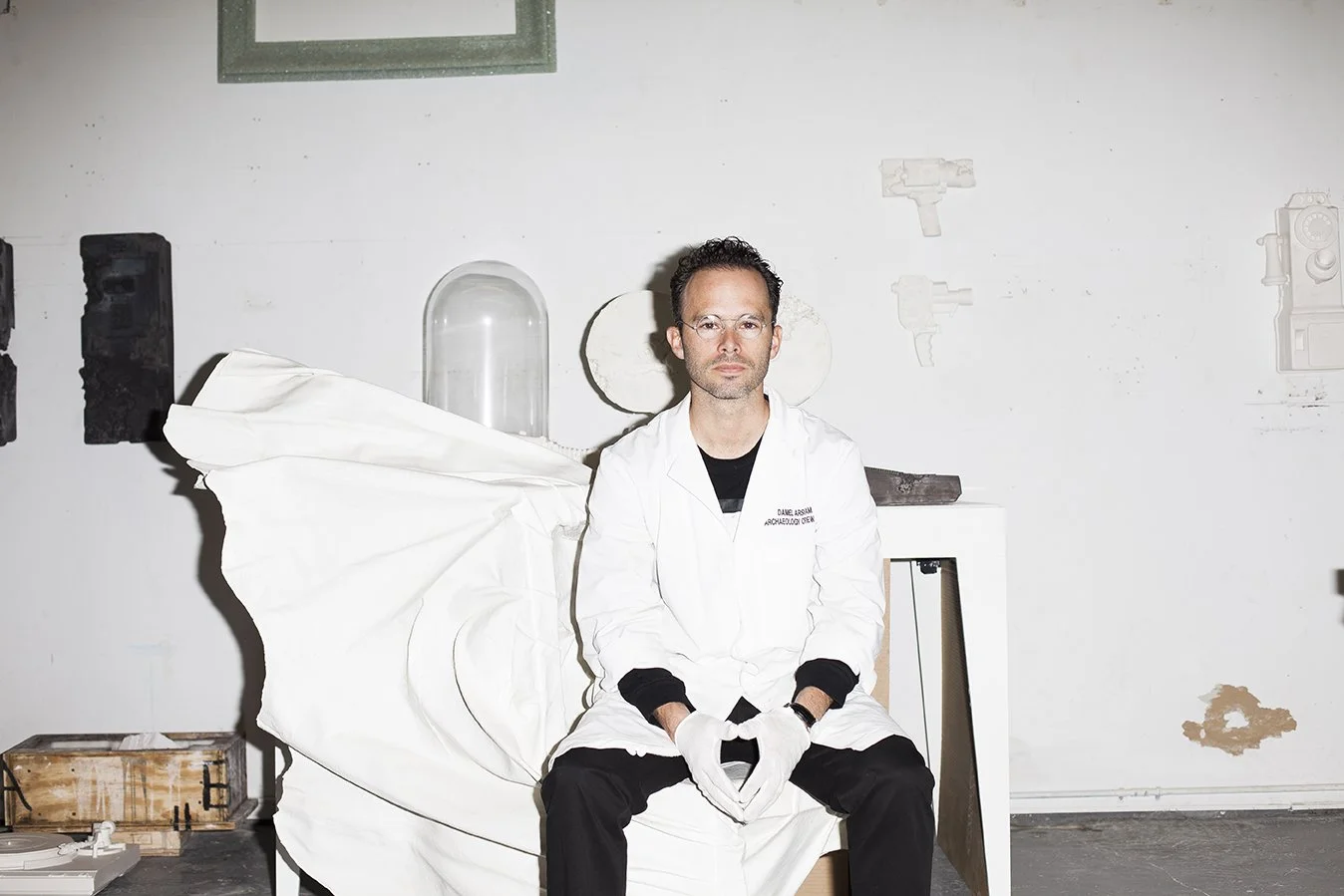 A man sits on a white examination table in a minimalist room. He wears a white coat with black pants, gloves, and glasses. The background features a white wall with faint outlines of various objects, and some worn spots on the wall.