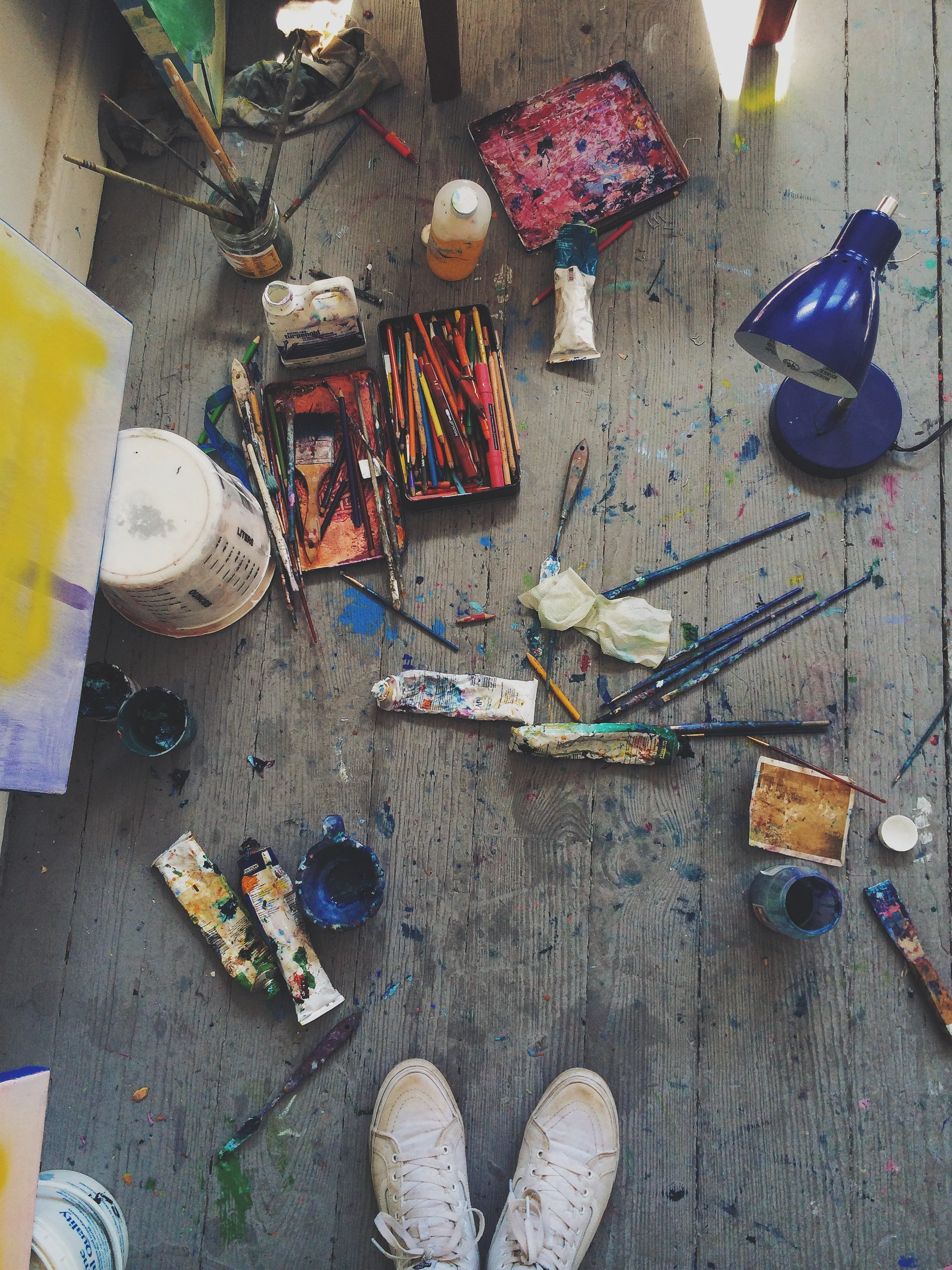 Overhead view of an artist's messy workspace on a wooden floor, with paint tubes, brushes, a palette, and a desk lamp, as well as a person's feet in white sneakers at the bottom.