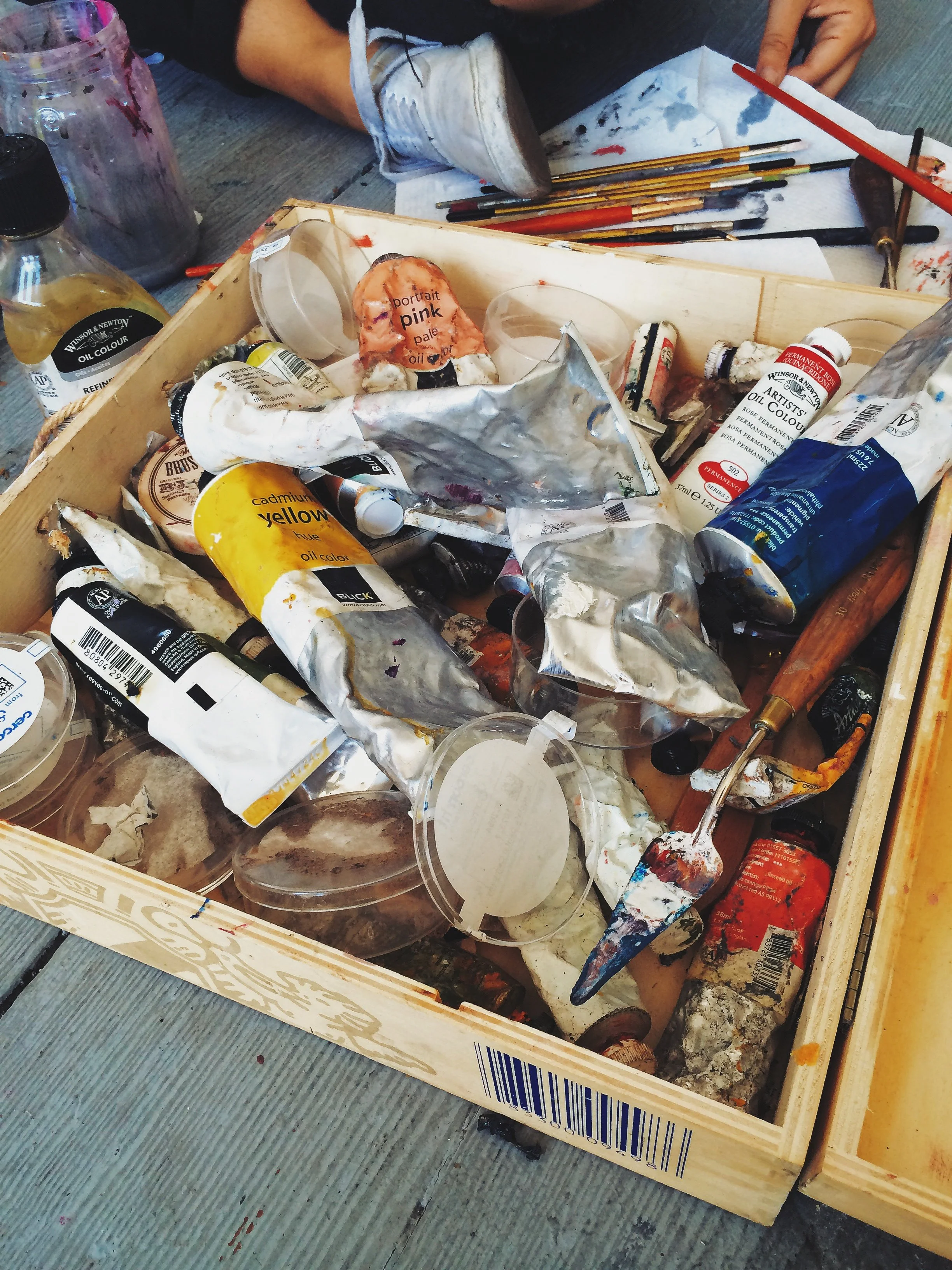 A wooden art palette box filled with used oil paint tubes, some squeezed and crumpled, along with painter's tools such as brushes, a palette knife, and a small jar, with a person sitting nearby on a wooden surface.