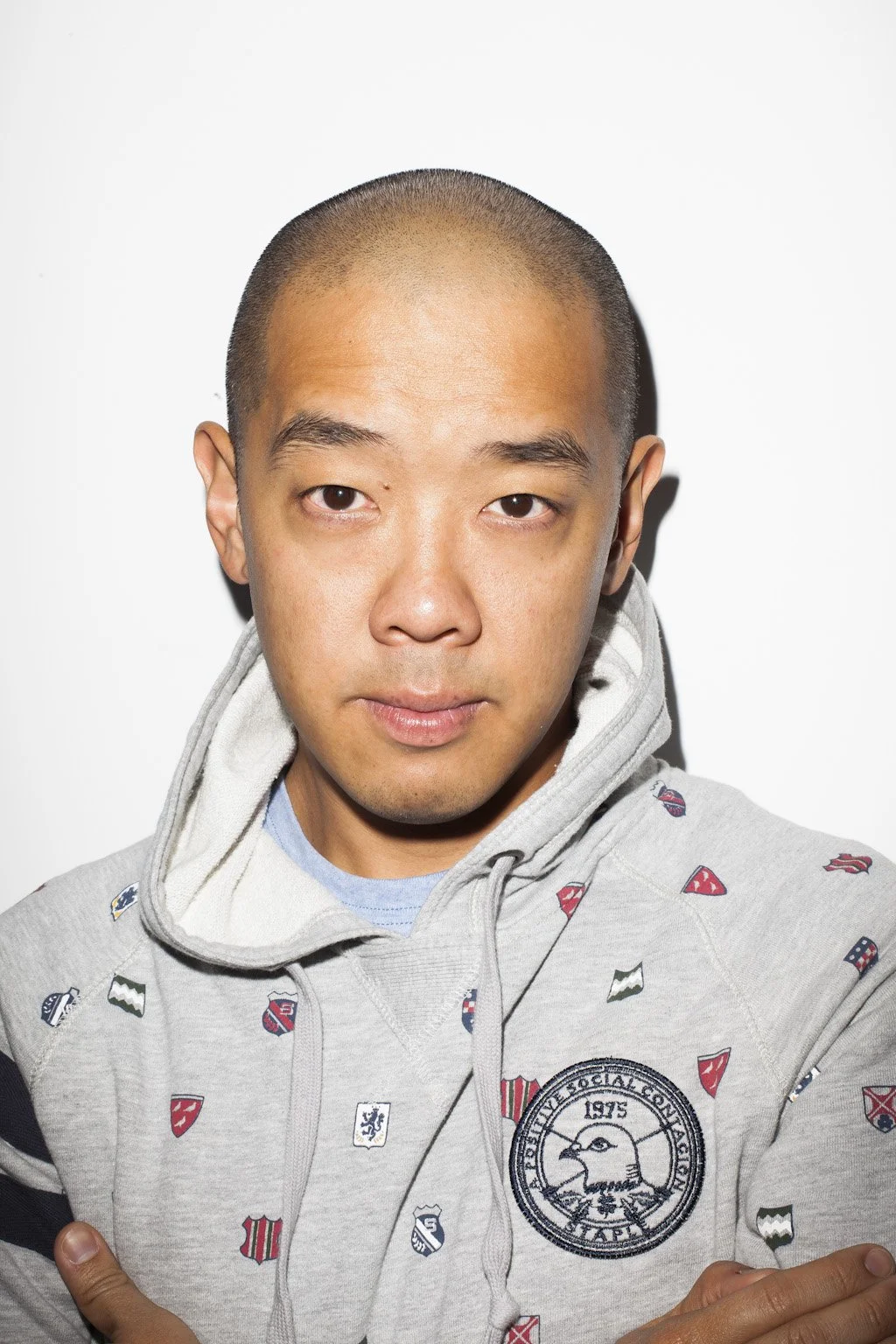 A young man with a shaved head wearing a gray hoodie with various patches and designs, standing against a plain white background.