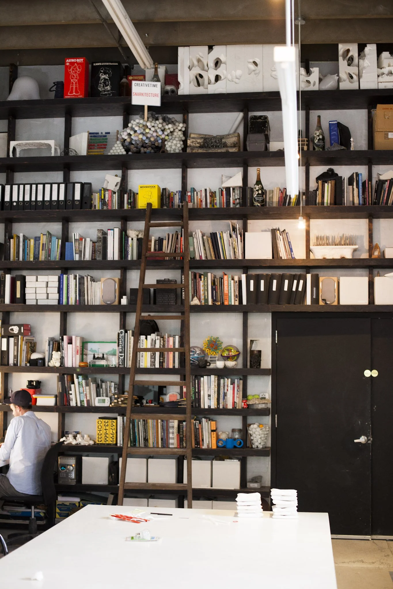 A large wall-mounted bookshelf filled with books, decorative objects, and storage boxes in an industrial-style room. A wooden ladder leans against the shelves for access.