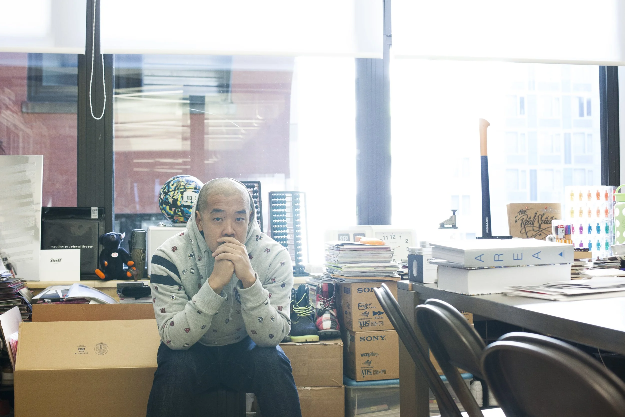 A man sitting in an office full of clutter, wearing a gray hoodie with patterns, with his hands clasped in front of his face, looking serious.