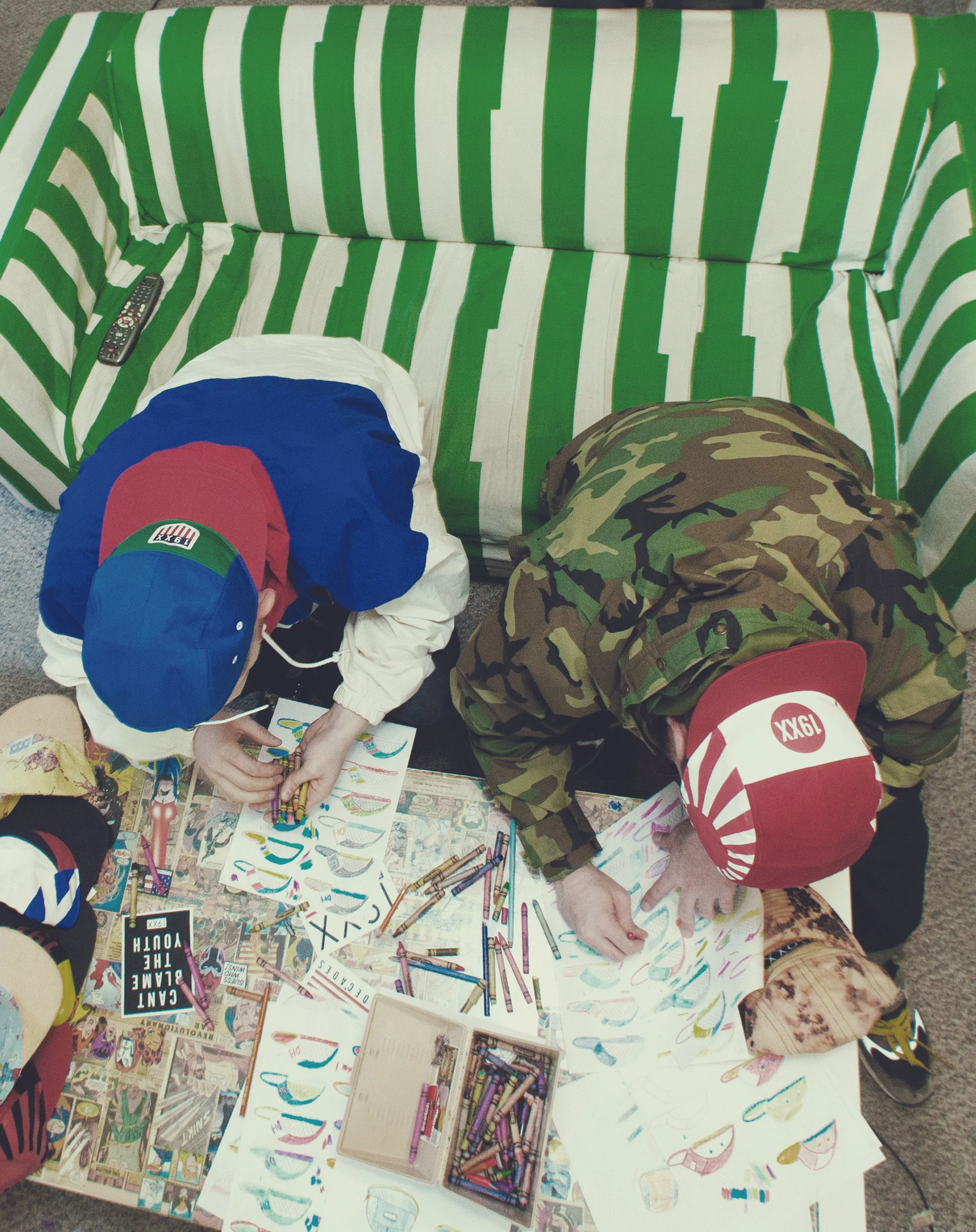 Two children wearing red and blue hats and camouflage clothing are drawing and coloring paper fish illustrations on a table covered with comic book pages, crayons, and markers.