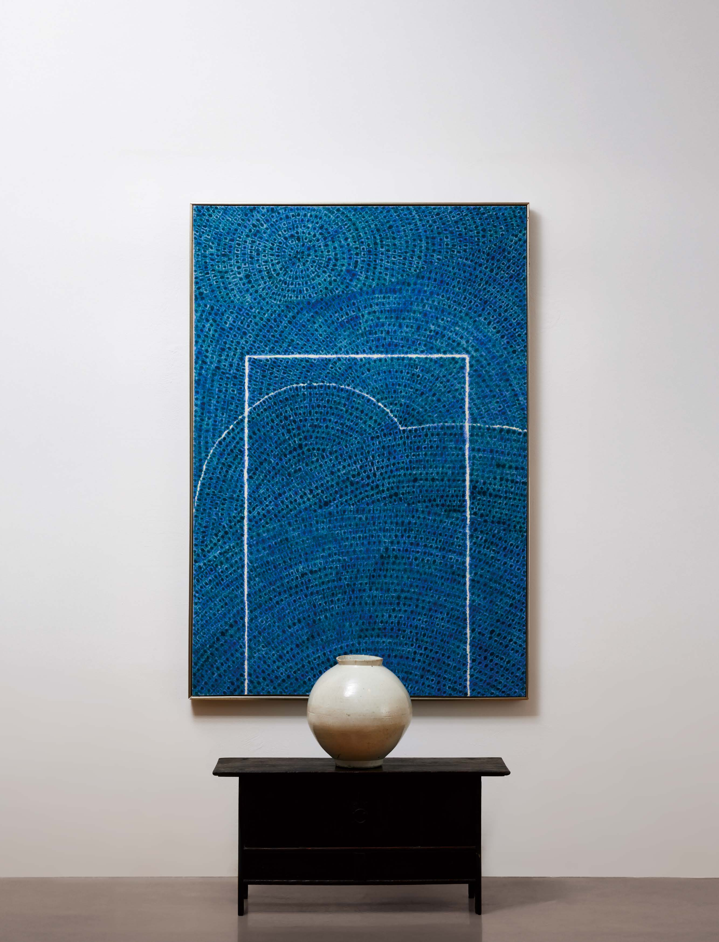 A white vase on a dark wooden table in front of an abstract blue artwork with geometric patterns.