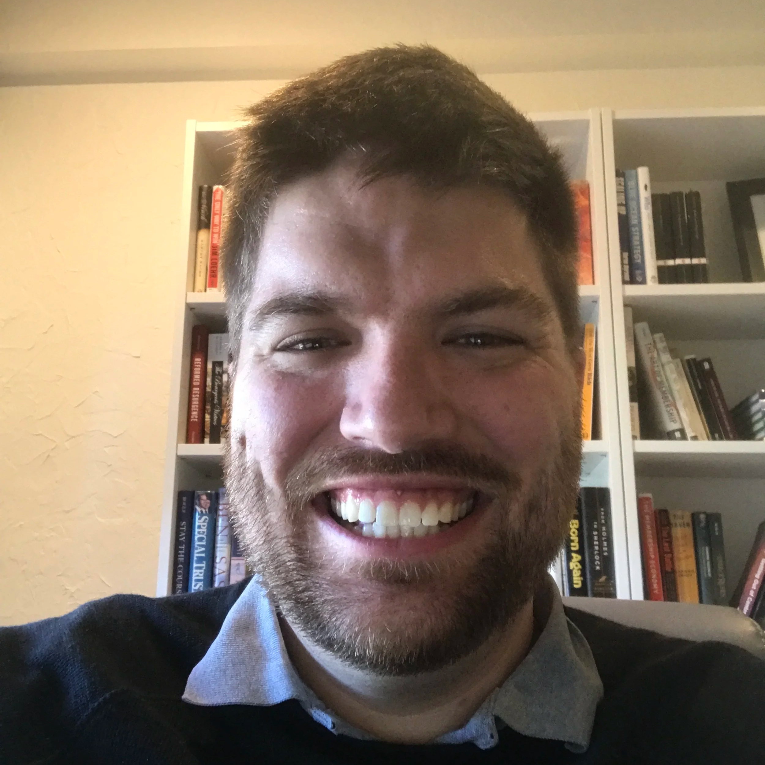 A man with short brown hair and a beard smiling showing his teeth, wearing a dark sweater over a collared shirt, in front of a bookshelf filled with books.