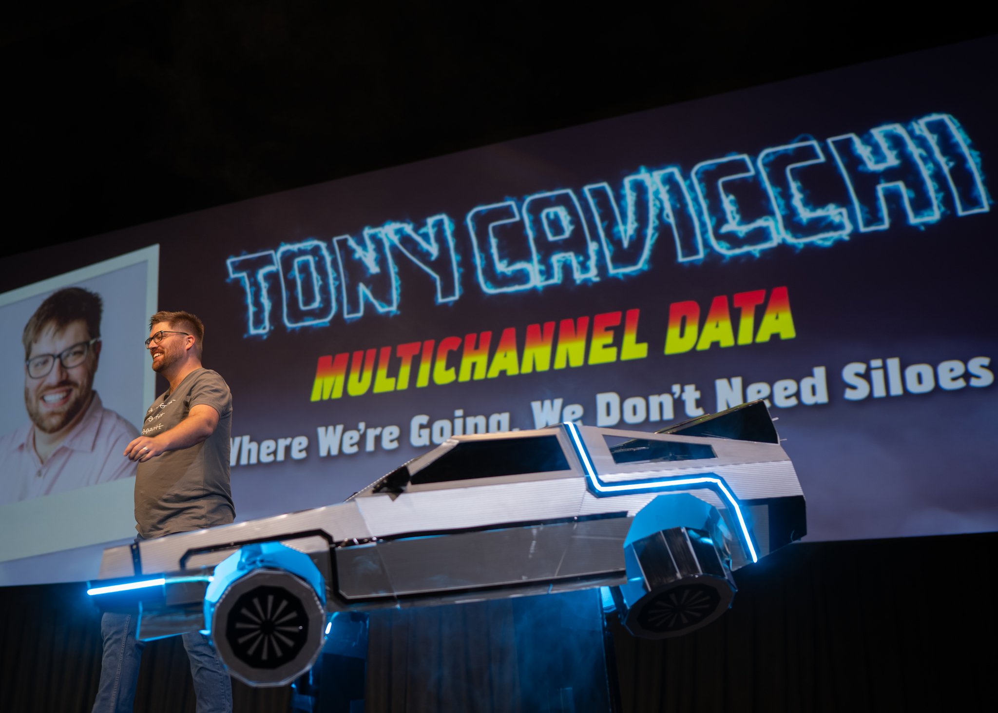Man on stage smiling, standing next to a futuristic vehicle with neon accents, in front of a large screen displaying a presentation with the title 'TONY CAVICCHI' and text about multichannel data.