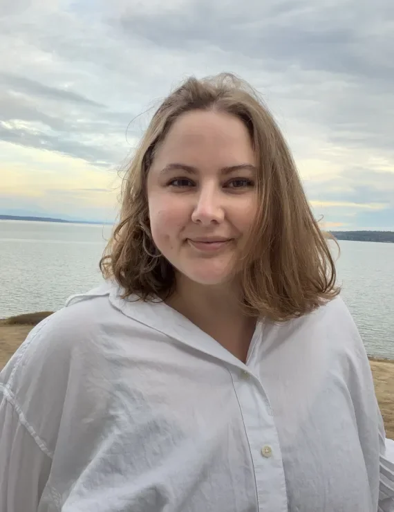 A nonbinary therapist named Audrey Meadowcroft, LMHCA, with shoulder-length light brown hair is smiling, standing outdoors near the Putget Sound in Seattle, Washington near their office in Fremont.