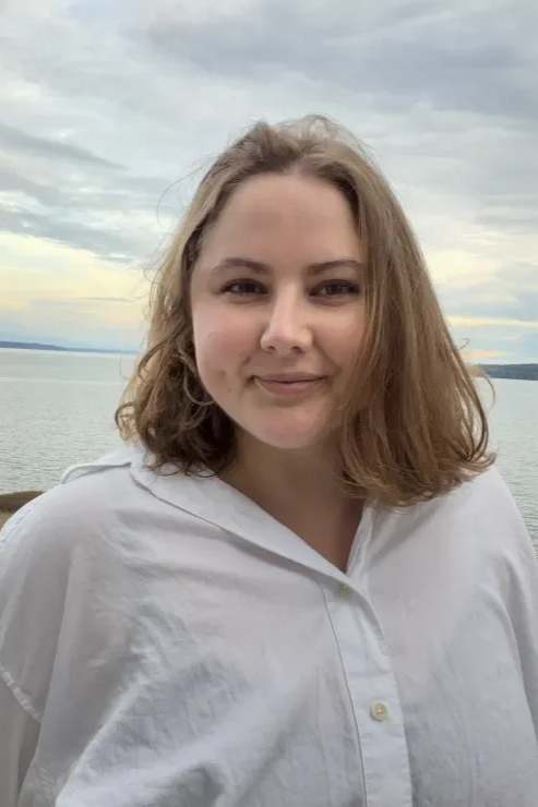 A young woman with wavy, shoulder-length hair outdoors near a body of water with a cloudy sky in the background.