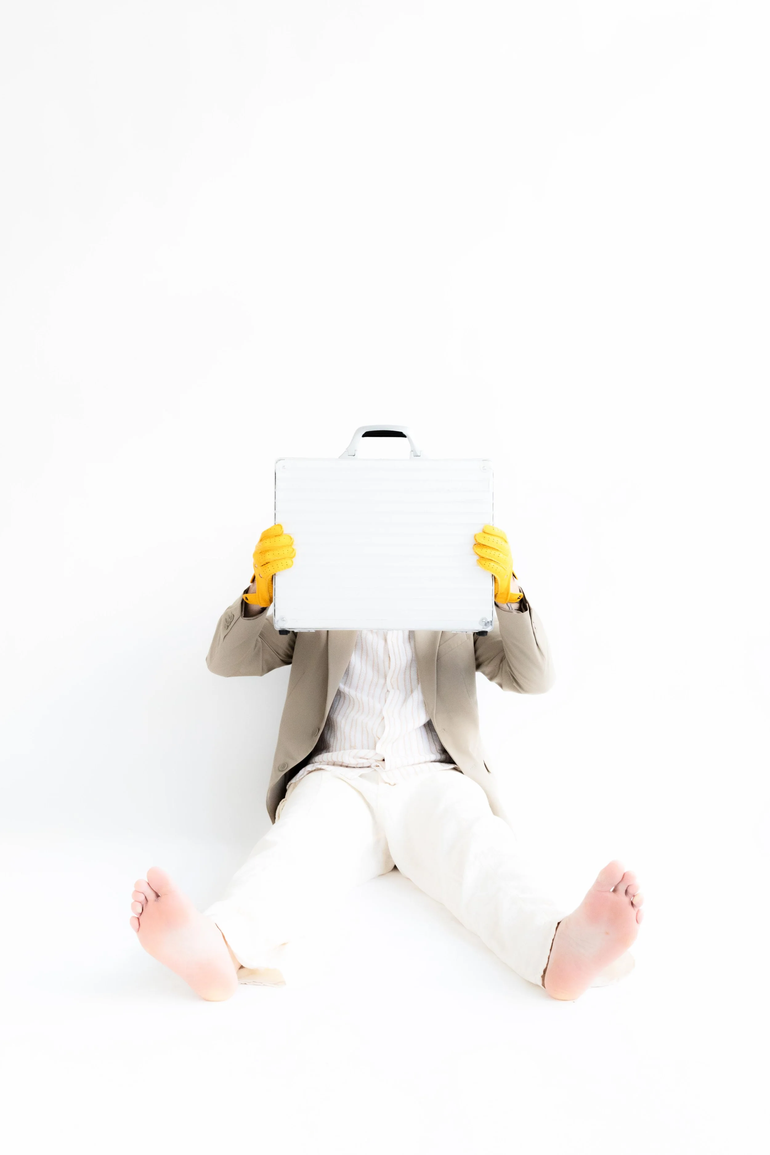 Person sitting barefoot on the floor, wearing a beige jacket and pants, holding a metal briefcase in front of their face, with yellow gloves on their hands, against a white background.