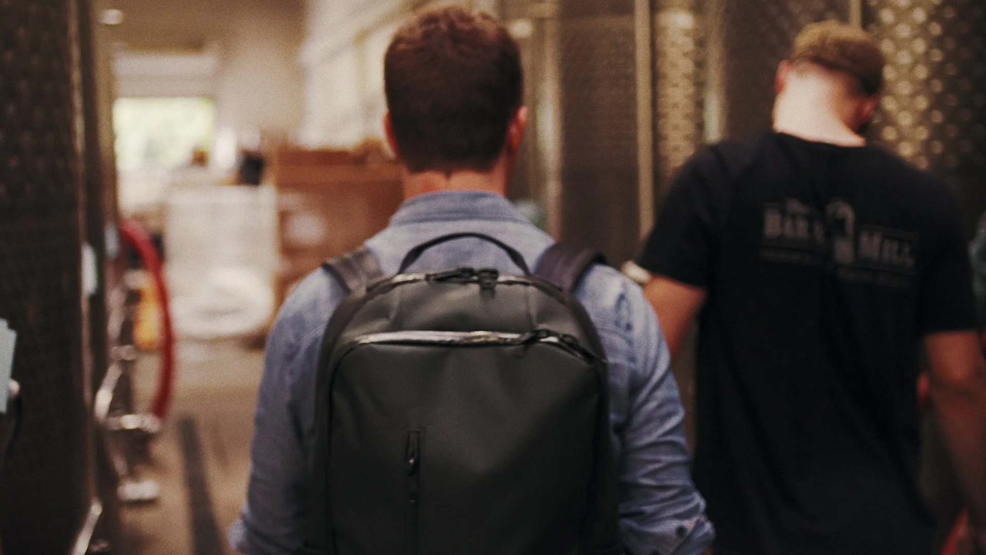 Two young men walking in a hallway, facing away from the camera. One is wearing a gray shirt and carrying a black backpack, while the other is wearing a black shirt with text on the back.