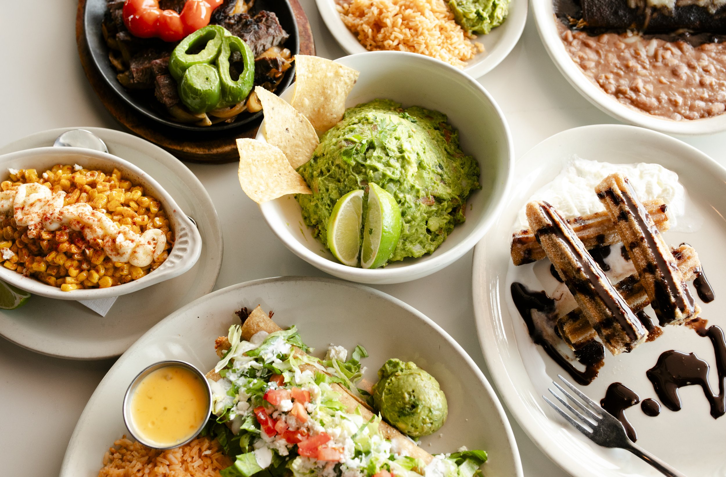 A variety of Mexican dishes including guacamole with lime wedges and tortilla chips, grilled meat with peppers, Mexican rice, refried beans, corn salad, and churros with chocolate sauce for dessert.