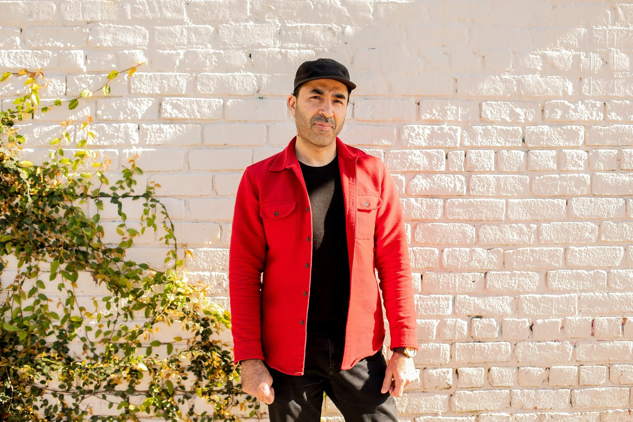 A man wearing a black cap, red jacket, and black shirt standing outdoors in front of a white brick wall with a green vine on the left.