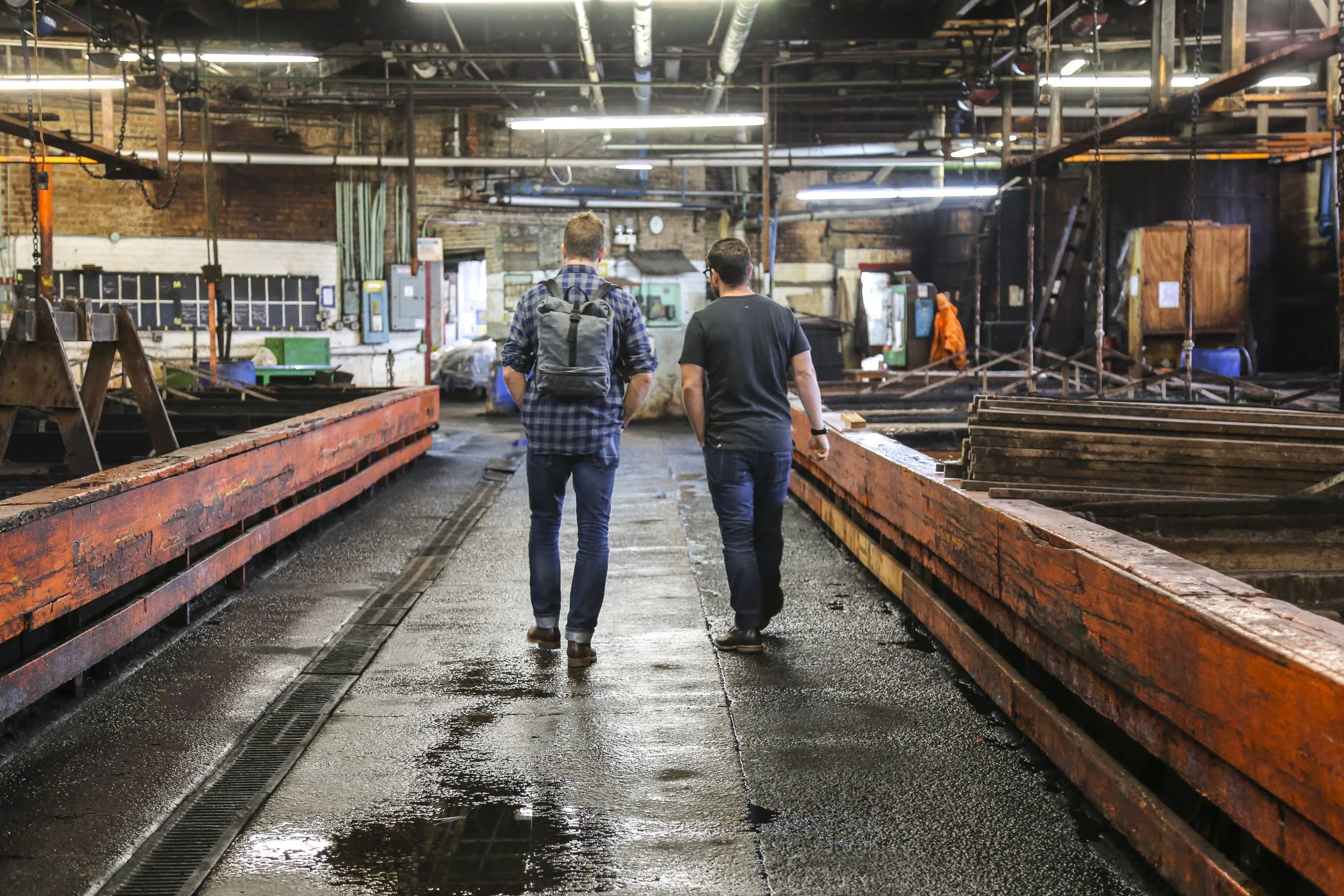 Two men walk through an industrial workshop or factory, surrounded by machinery, wooden beams, and metal structures, with wet floors and overhead lighting.