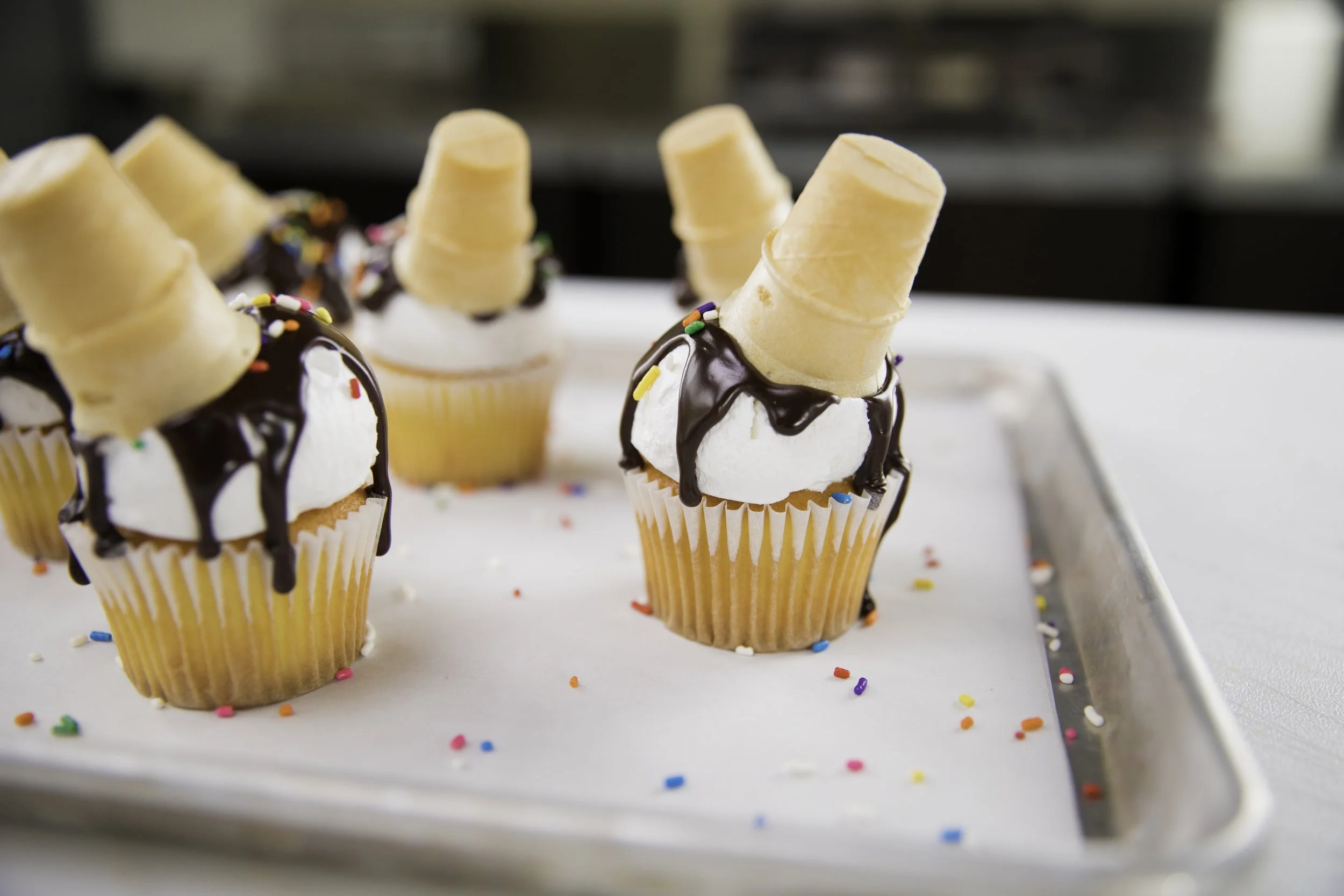 Cupcakes topped with white frosting, chocolate drizzle, colorful sprinkles, and mini ice cream cones, on a metal tray with scattered sprinkles.