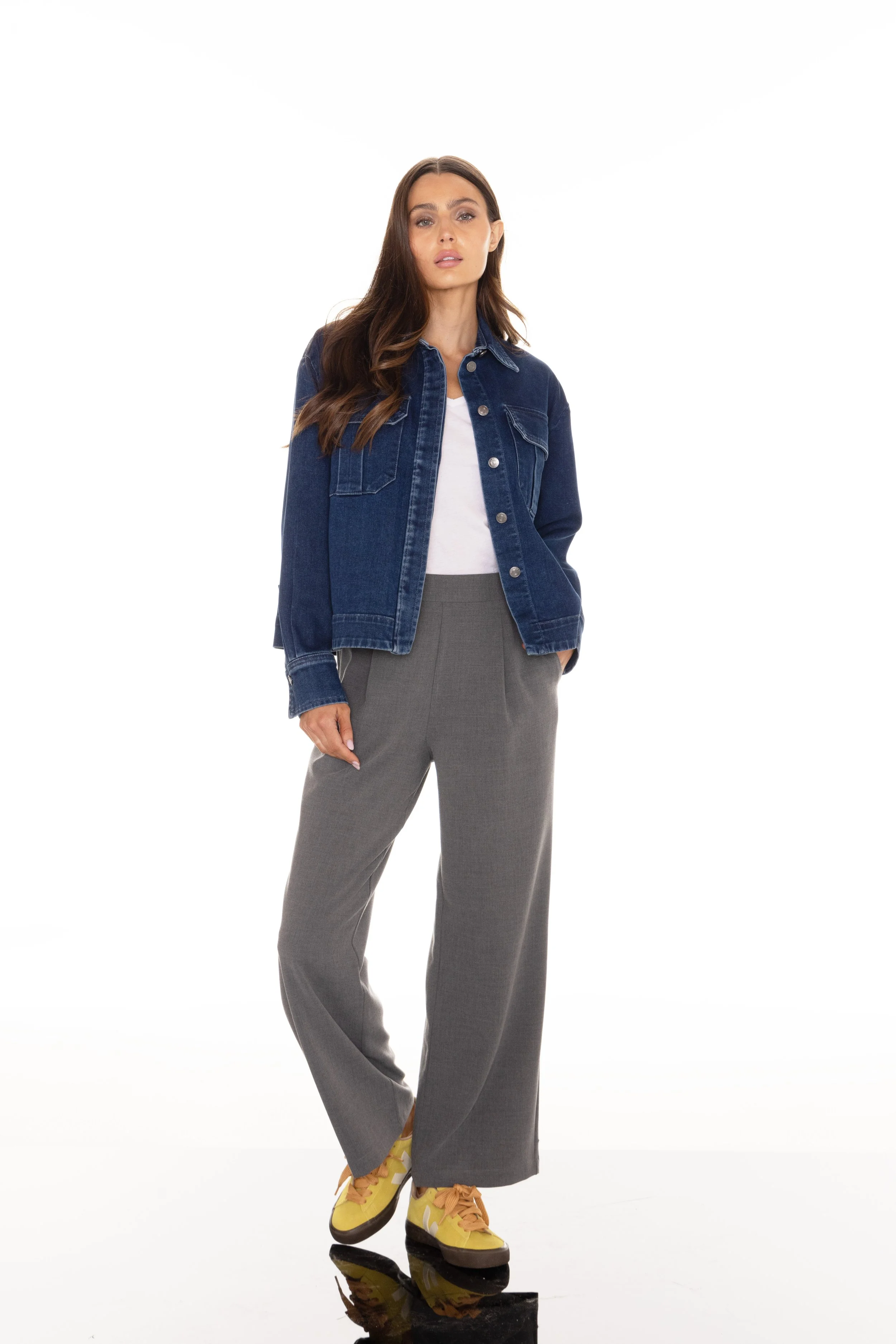 Woman standing with hands in pockets, wearing a denim jacket, gray wide-leg pants, white t-shirt, and yellow sneakers on a white background.