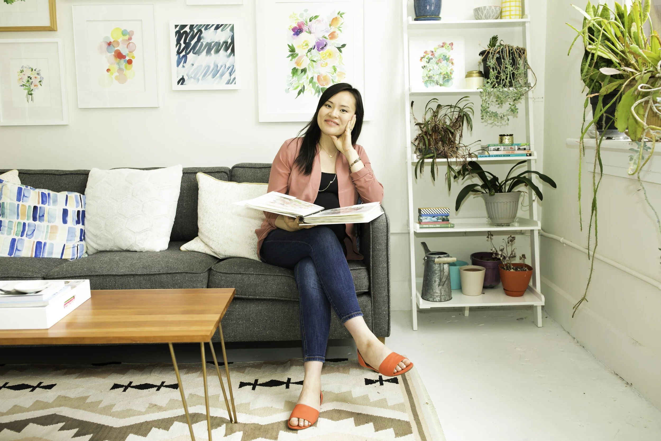 Yeo Cheng is sitting on a gray sofa in a living room, holding open a photo album, smiling, with art prints on the wall behind her and a white shelf filled with plants and books to her right.