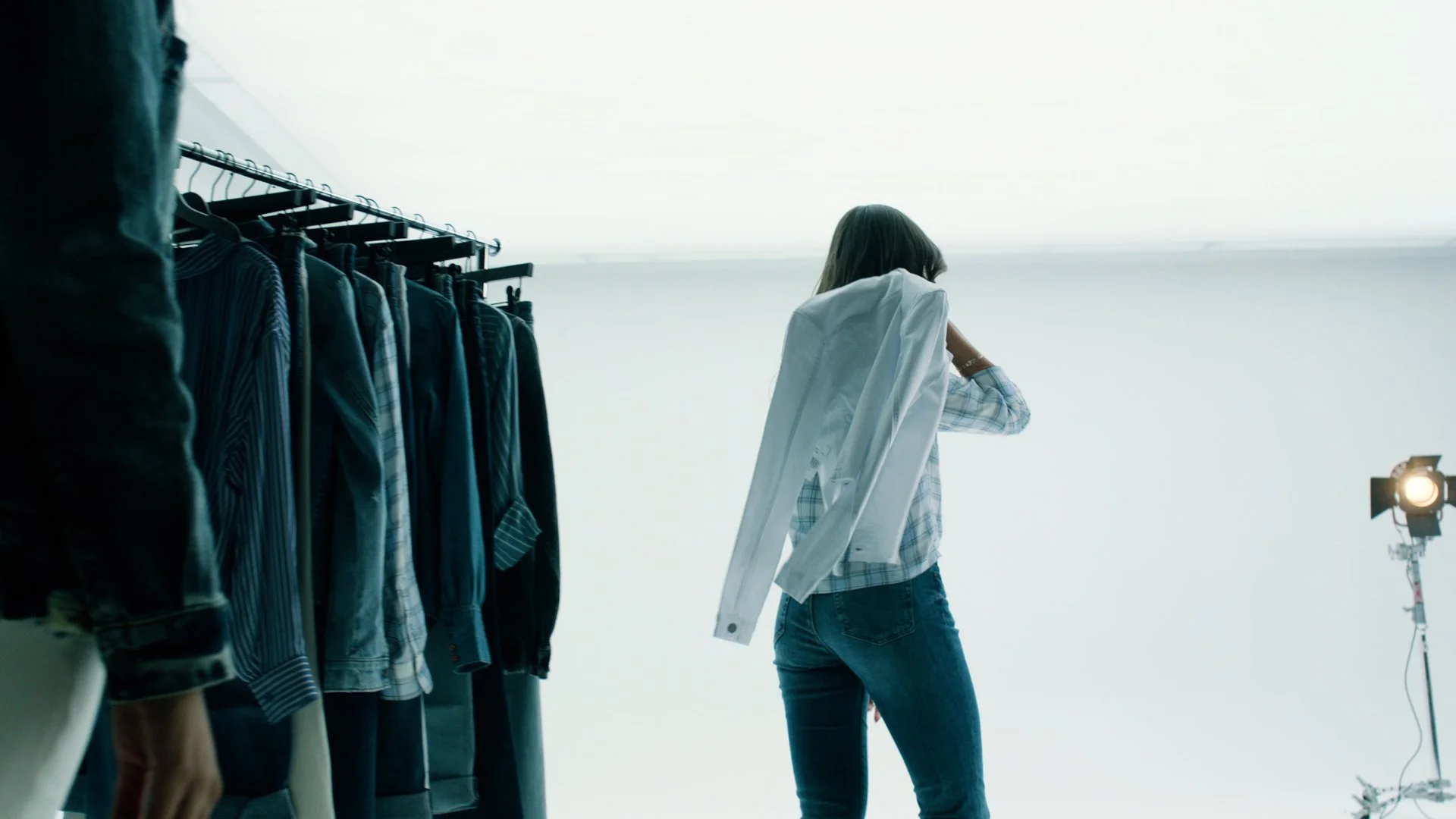 A woman standing in front of a clothing rack filled with various shirts, wearing a plaid shirt, jeans, and holding a white jacket over her shoulder. A professional studio light is visible on the right side of the image.