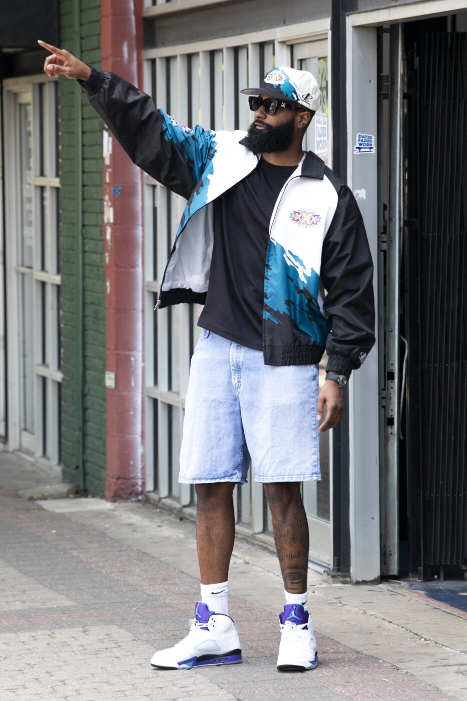 A man standing on a city sidewalk, pointing to his left. He's wearing sunglasses, a white and blue patterned cap, a black t-shirt, a black and blue windbreaker, light denim shorts, white socks, and white and purple sneakers.