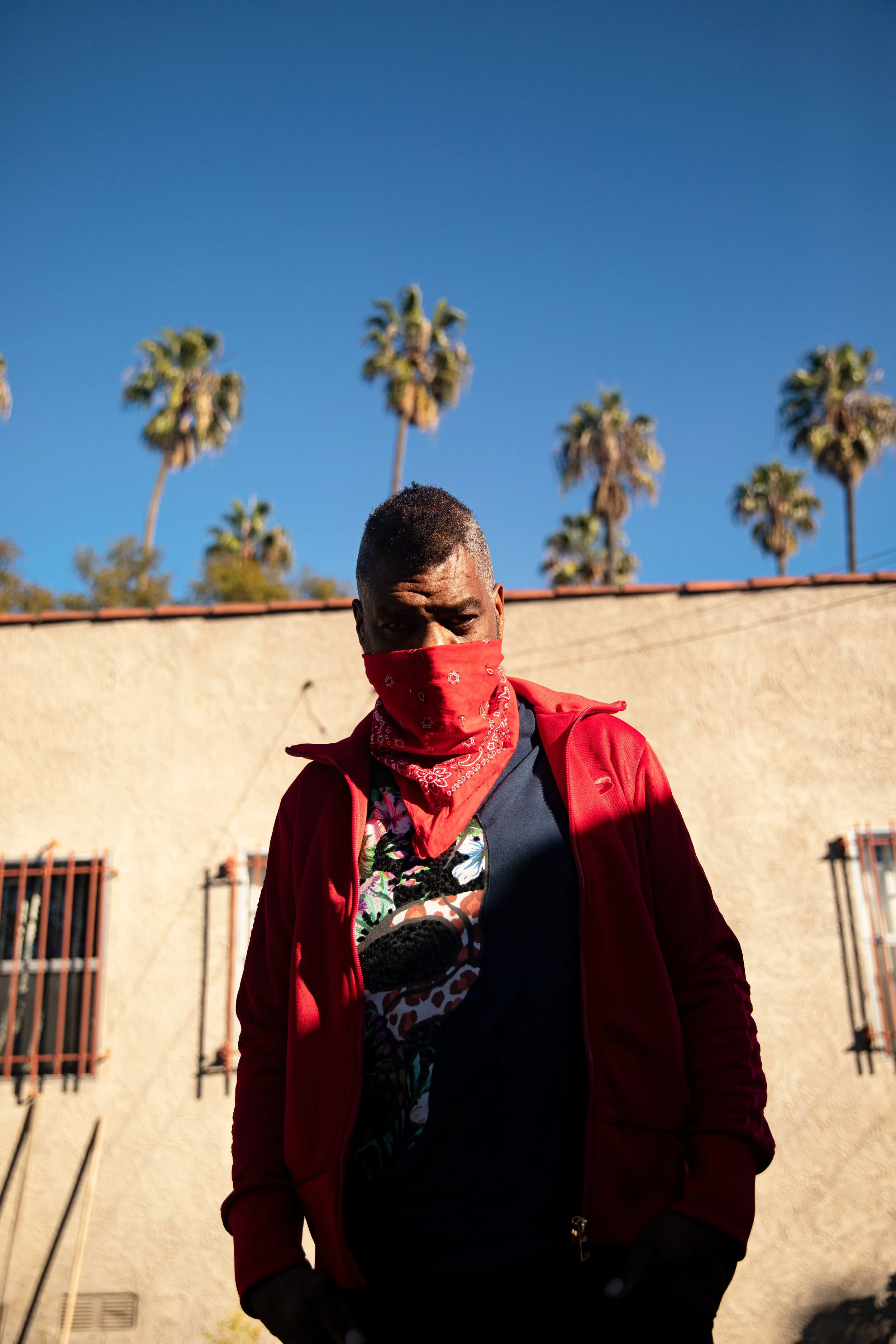 A man wearing a red bandana mask and a red jacket stands outdoors, partially illuminated by sunlight, with palm trees and a beige building with barred windows in the background.