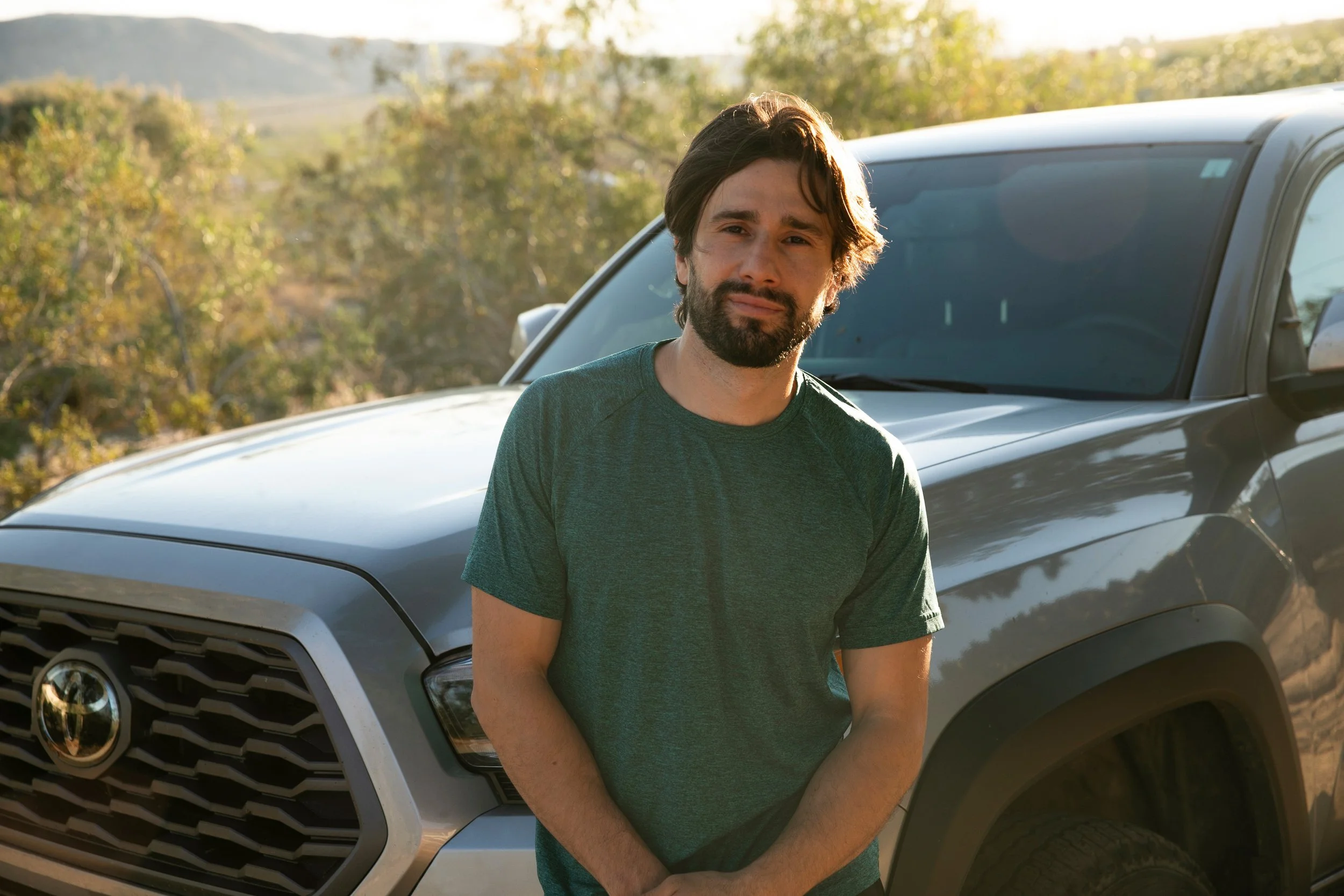 A man with dark hair and a beard standing outdoors in front of a silver Toyota SUV during daytime.