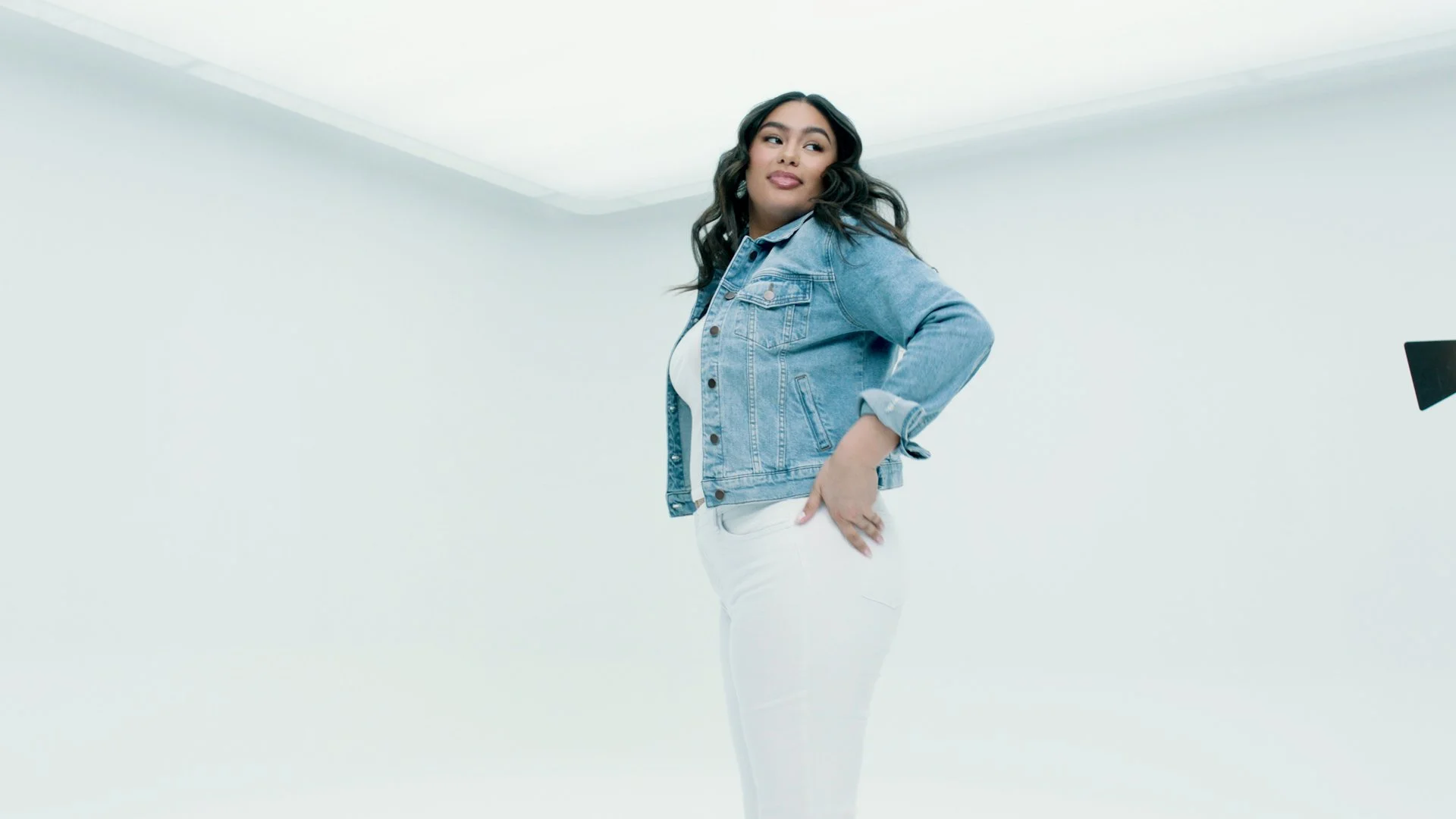 A young woman wearing a denim jacket and white pants posing with her hand on her hip in a well-lit room with plain white walls.