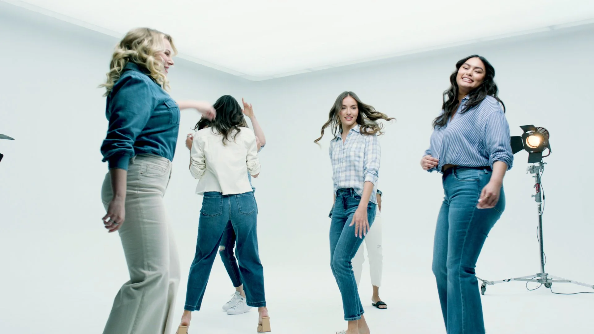 A group of five women dancing in a white studio, wearing casual jeans and shirts, with studio lighting equipment visible.