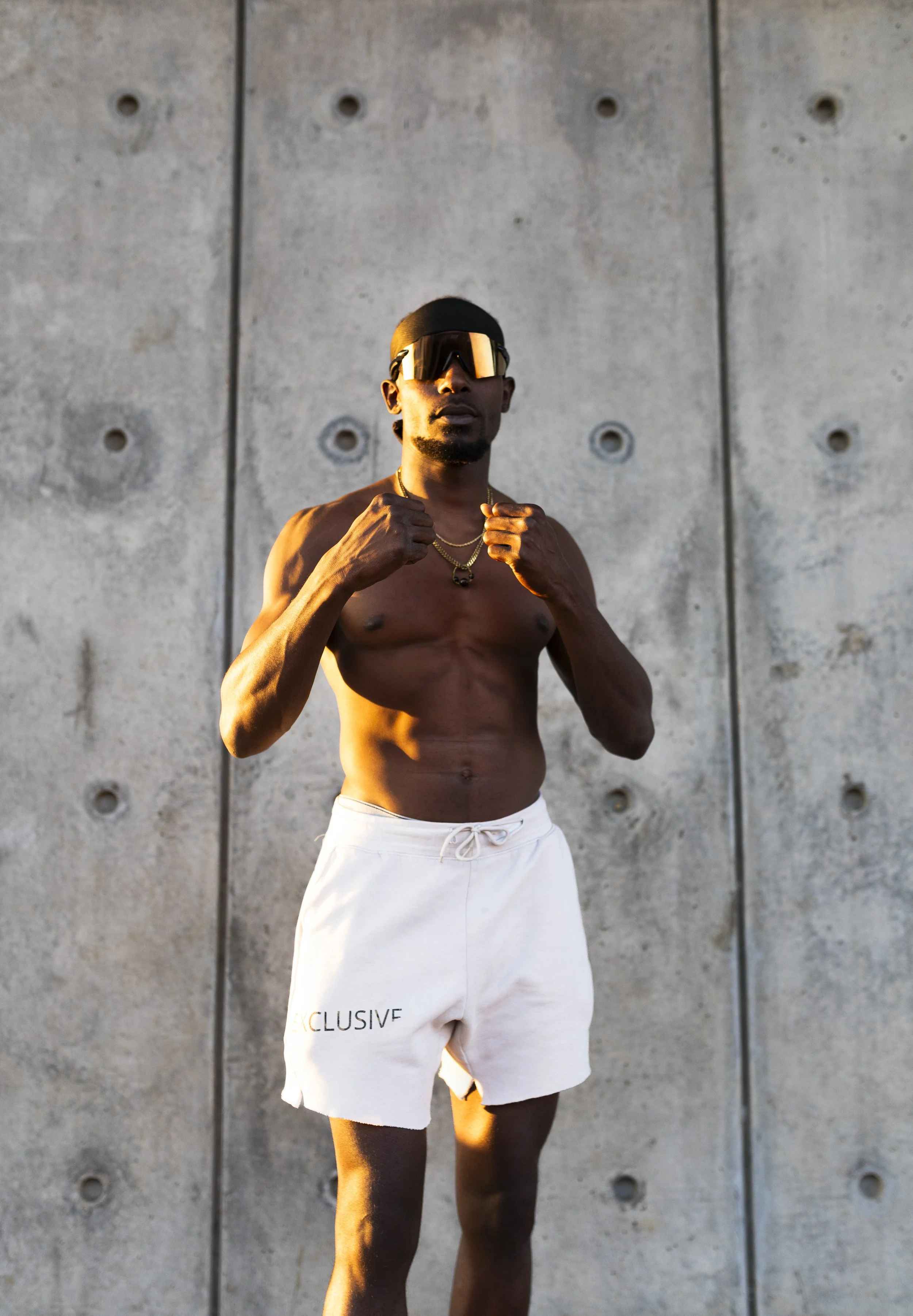 A shirtless man wearing sunglasses, necklaces, and white shorts with the word 'EXCLUSIVE' printed on them, standing in front of a concrete wall.
