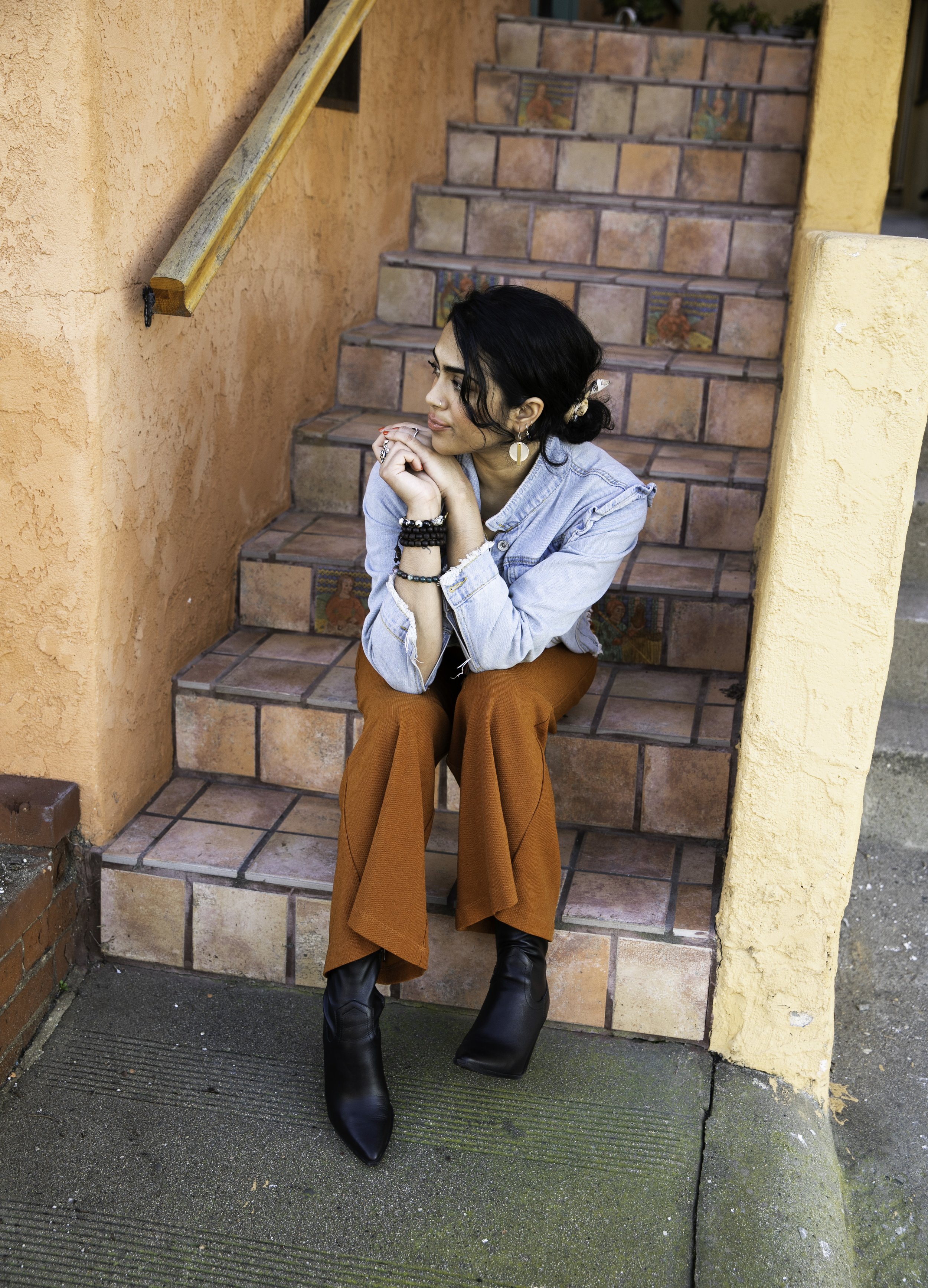 A woman with dark hair is sitting on outdoor tiled stairs with decorative tiles, resting her chin on her hand and looking to the side. She is wearing a light denim jacket, rust-colored wide-leg pants, black pointed boots, and multiple bracelets, with