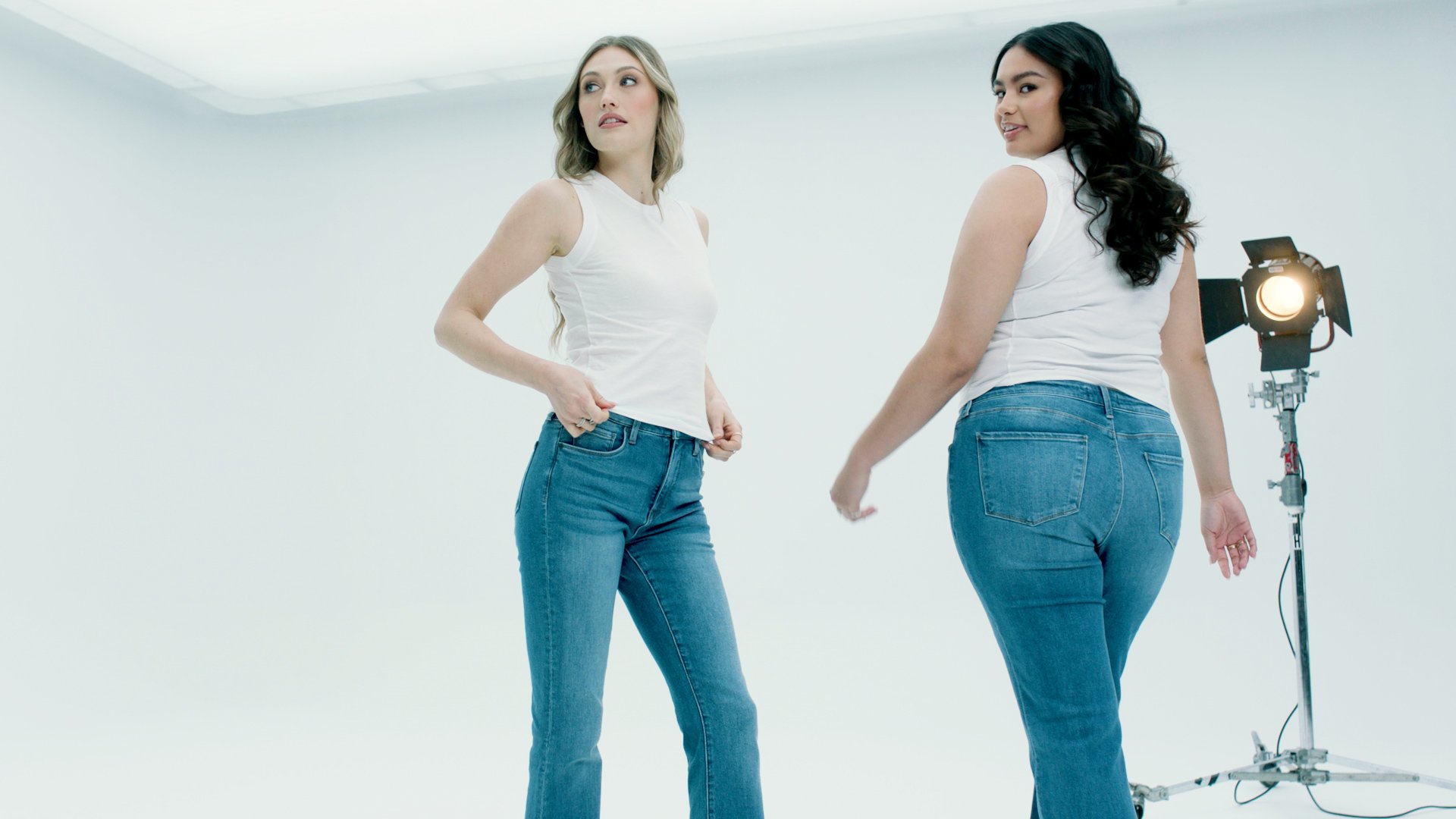 Two women in white tops and blue jeans standing in a photo studio with a professional light behind them.