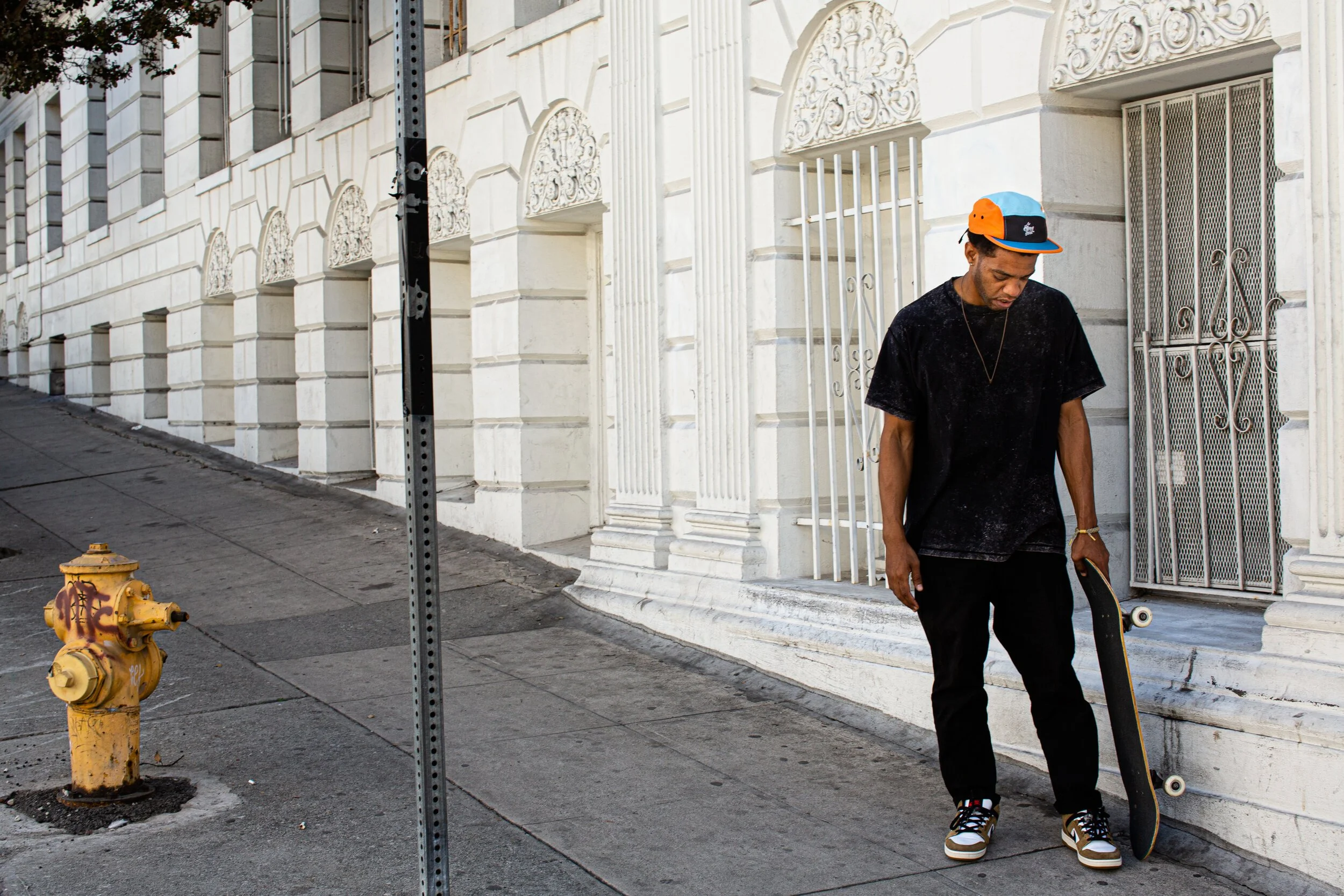 A man holding a skateboard standing on a city sidewalk next to a white building with ornate architectural details.