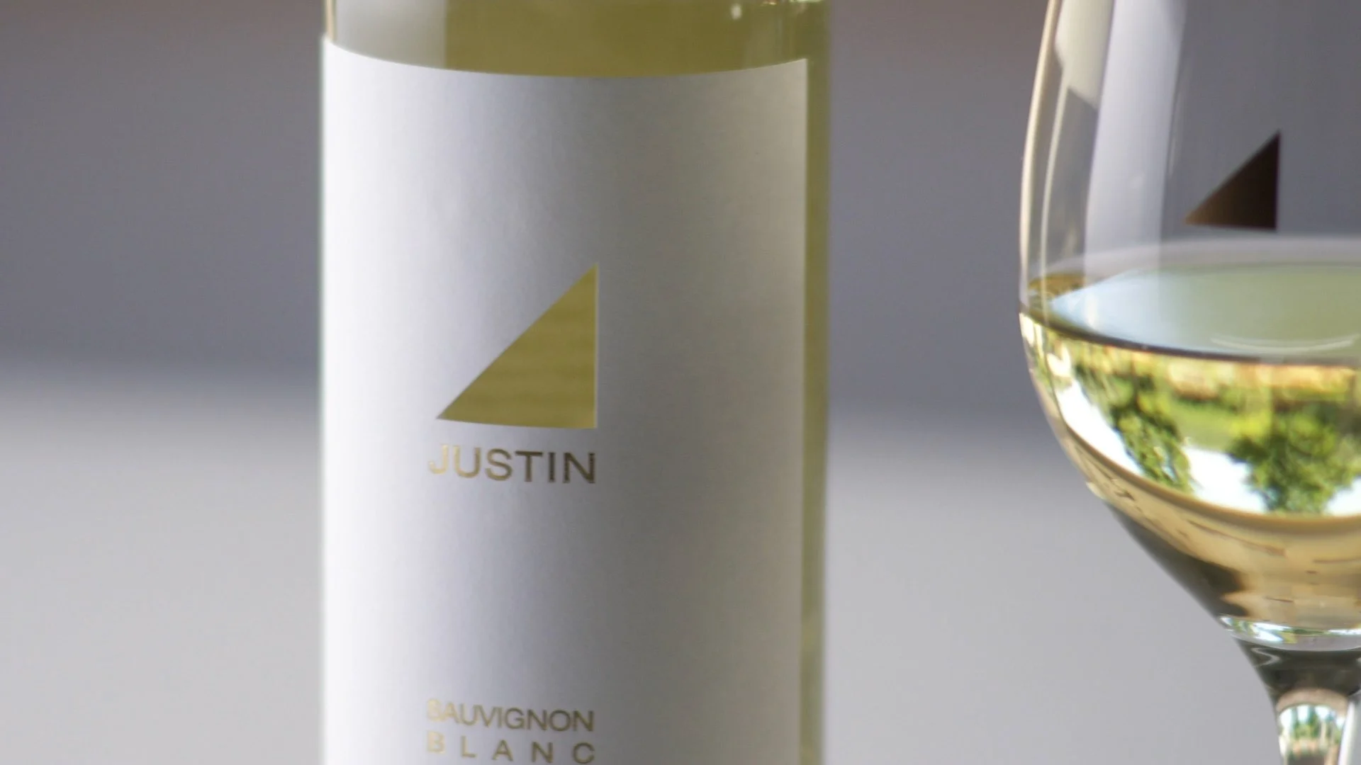 A bottle of Justin Sauvignon Blanc wine next to a filled wine glass, with a blurred outdoor reflection in the glass.