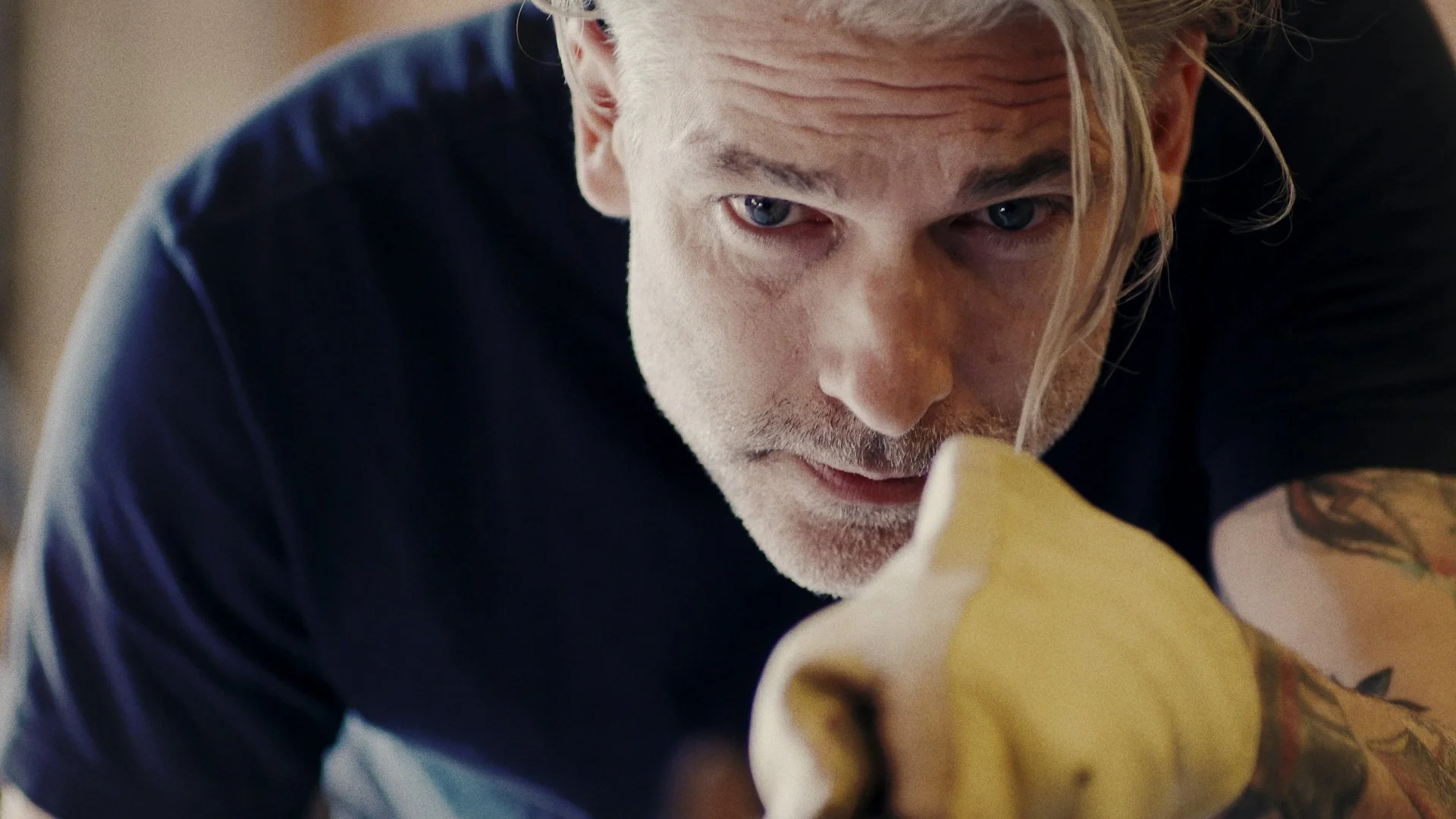 A man with blond hair and tattoos on his arm, wearing a black shirt and yellow gloves, is focused on a task.