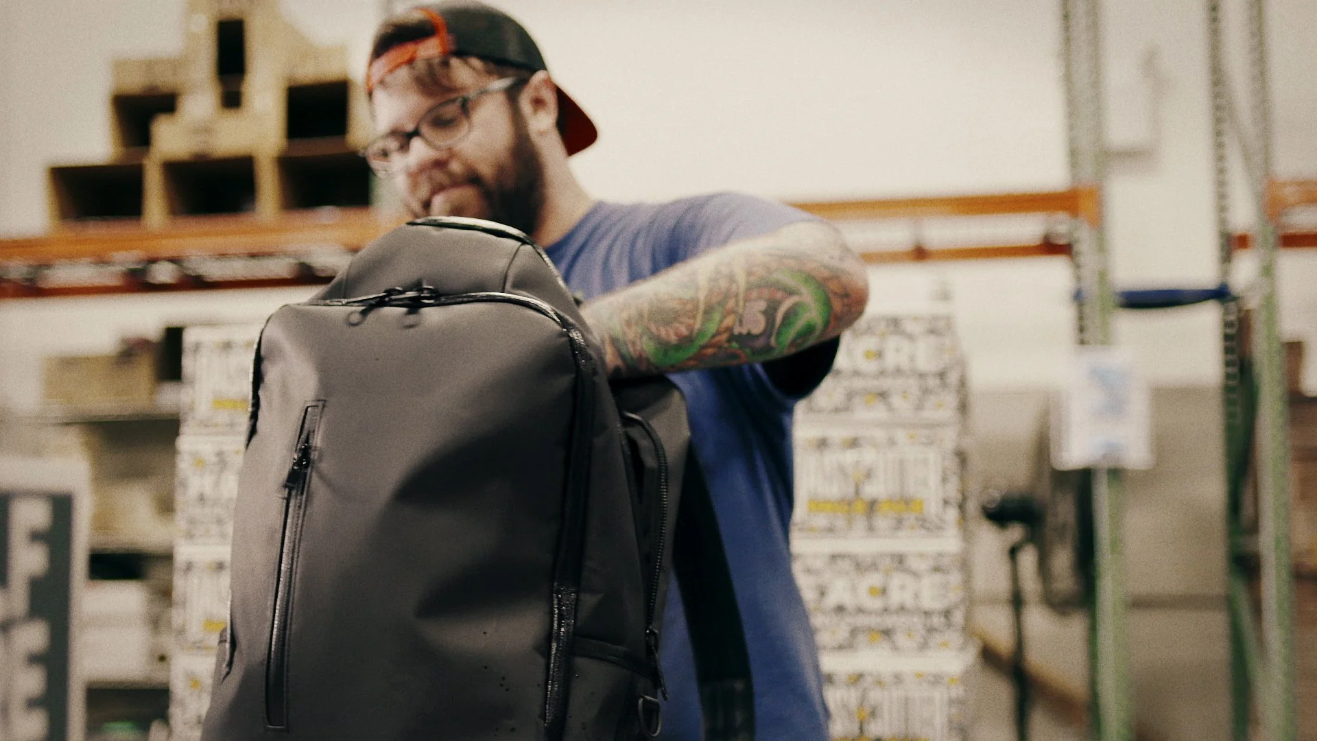 A man with glasses, a beard, and tattoos wearing a cap backwards is looking into a gray backpack in a warehouse or storage room.