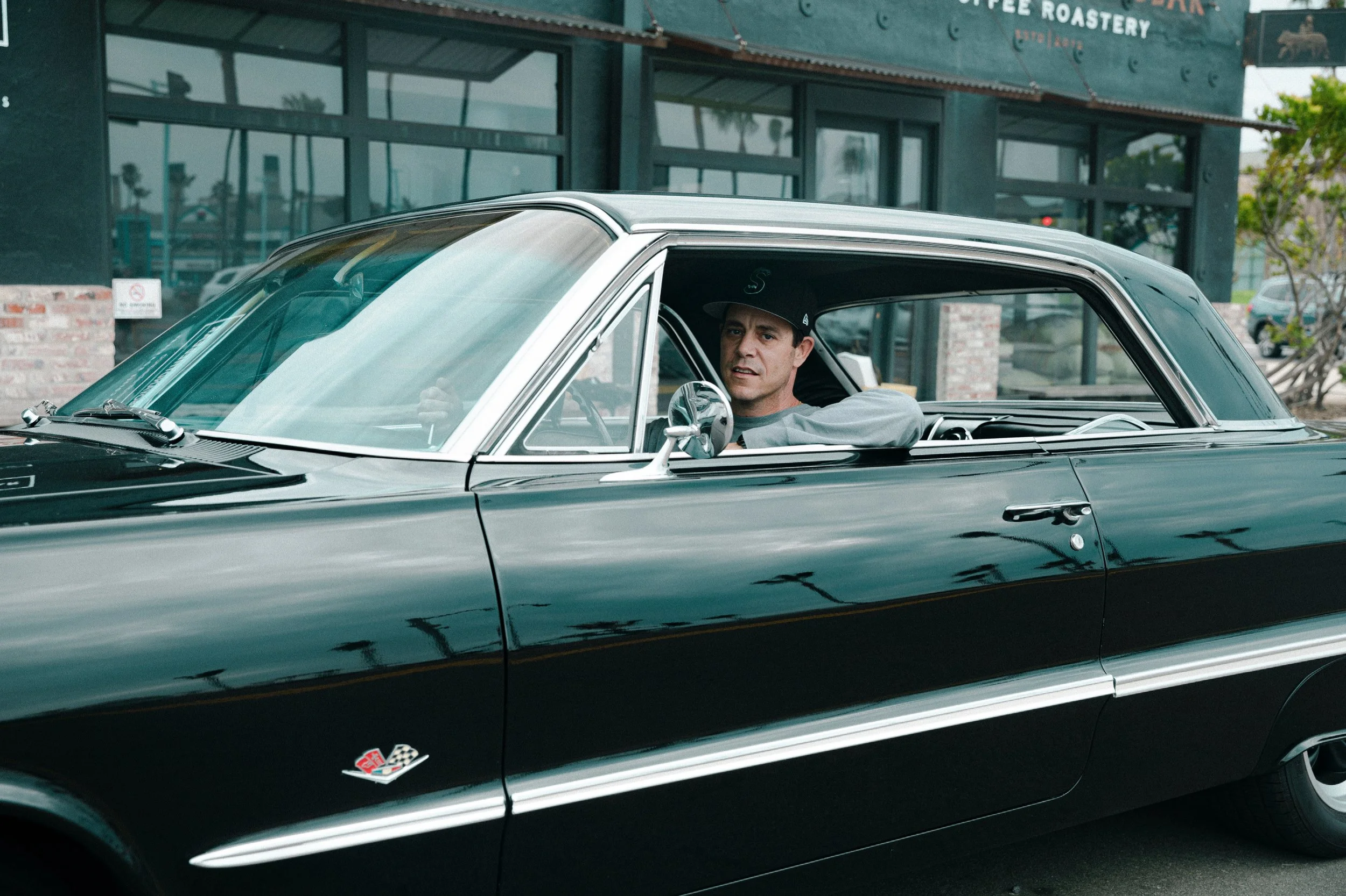 A man sitting in the driver's seat of a vintage black Chevrolet Corvette with a man wearing a black cap inside, outside a storefront with large windows and green signage.