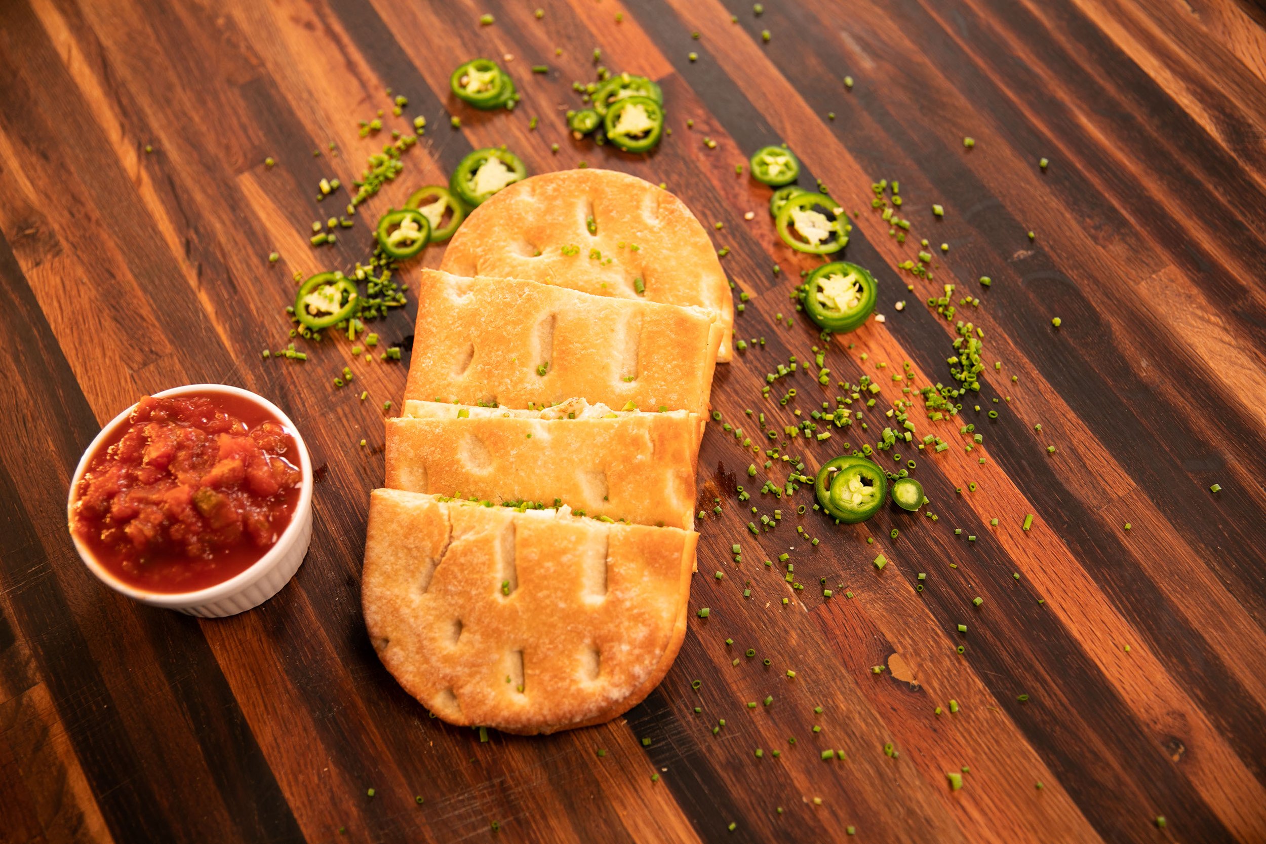 Four pieces of sliced flatbread with grill marks, chopped green jalapeños, and a small bowl of chunky salsa on a wooden surface.