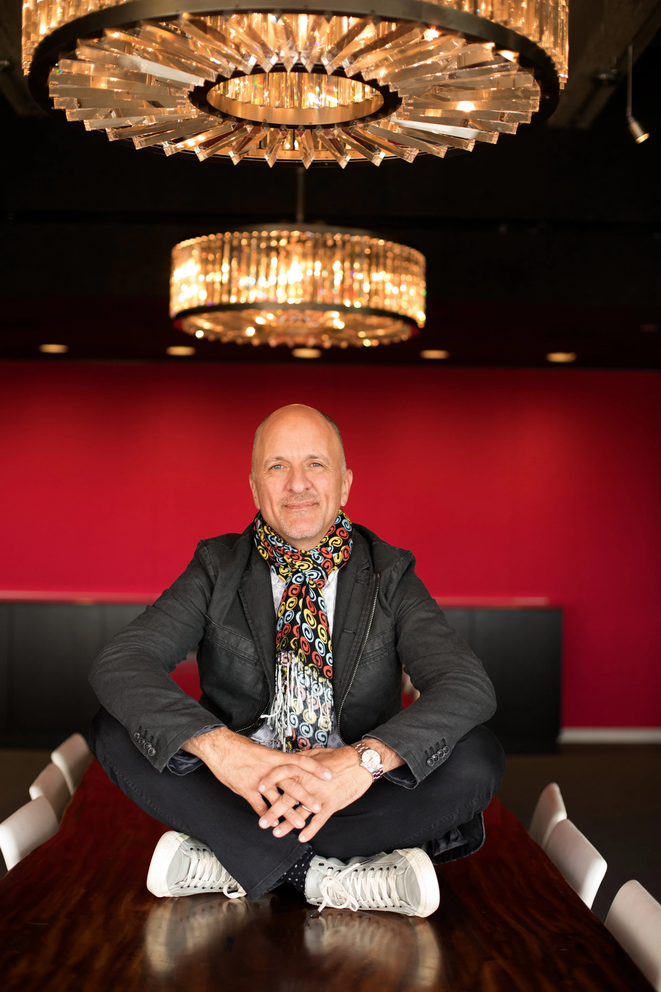 A man sitting cross-legged on a dining table, smiling at the camera inside a room with red walls and two large decorative chandeliers hanging from the ceiling.