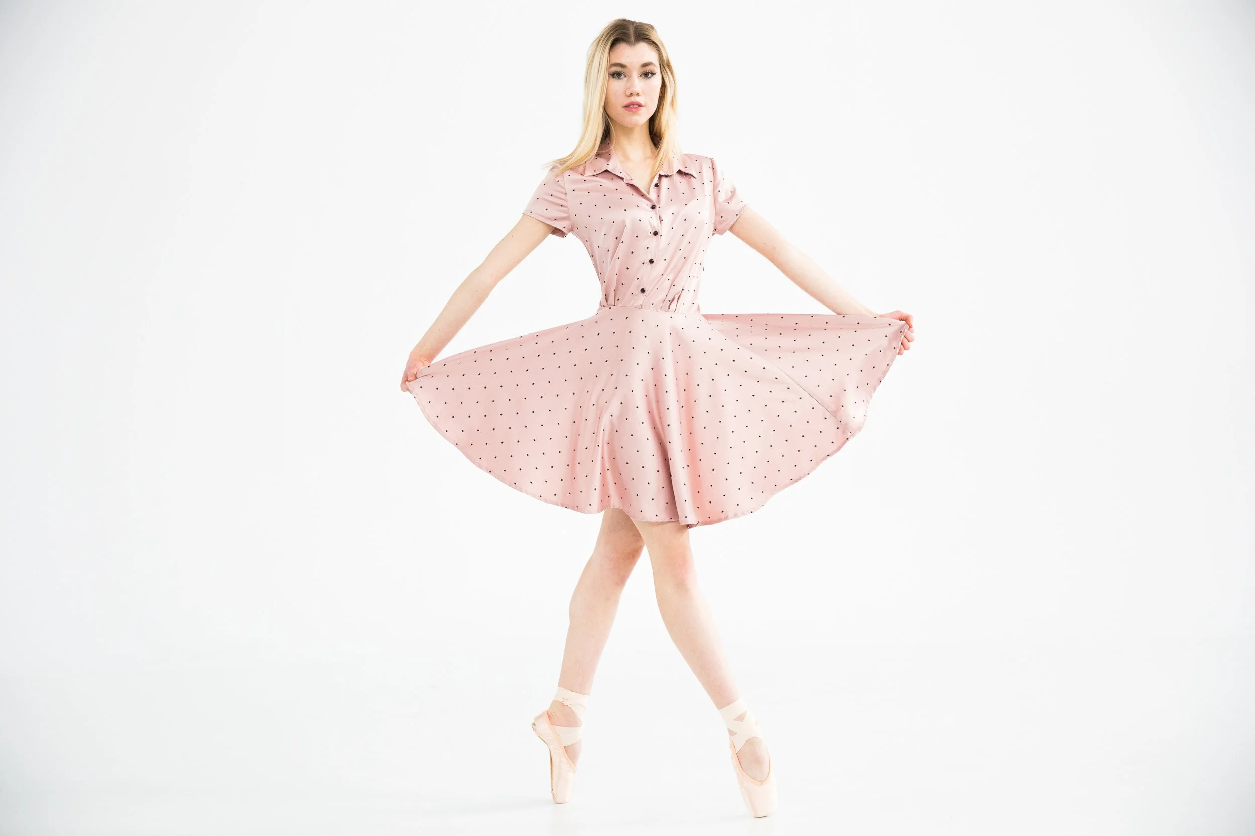 A young woman with blonde hair dressed in a pink polka-dot dress, standing on her toes in ballet pointe shoes, holding out the sides of her skirt against a plain white background.