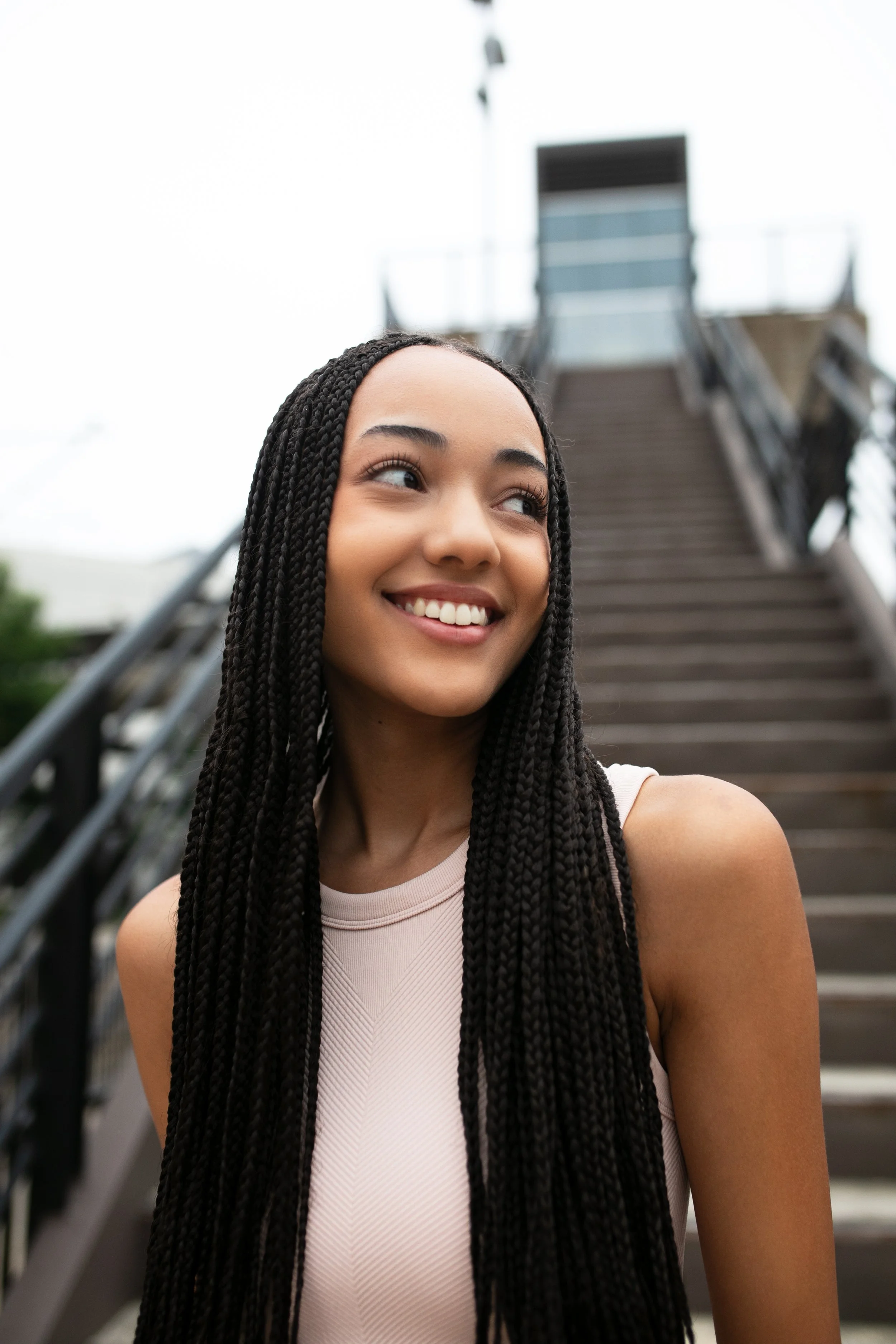A young woman with long braided hair smiling outdoors, with a staircase in the background.