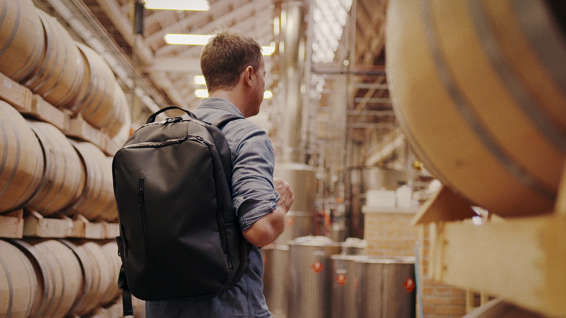 A man with a backpack standing in a brewery or winery, surrounded by large wooden fermentation tanks.