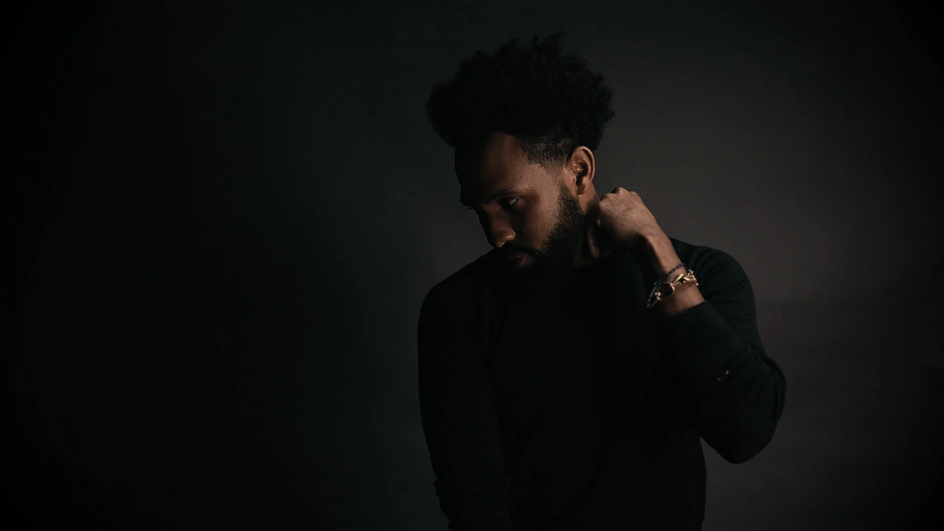 A dark-skinned man with curly hair and a beard wearing a black shirt, looking down and touching his neck with one hand in a low-lit setting.