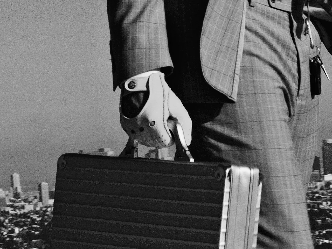 Partial view of a man in a plaid suit holding a briefcase in one hand and a white robotic hand in the other, with a cityscape in the background.